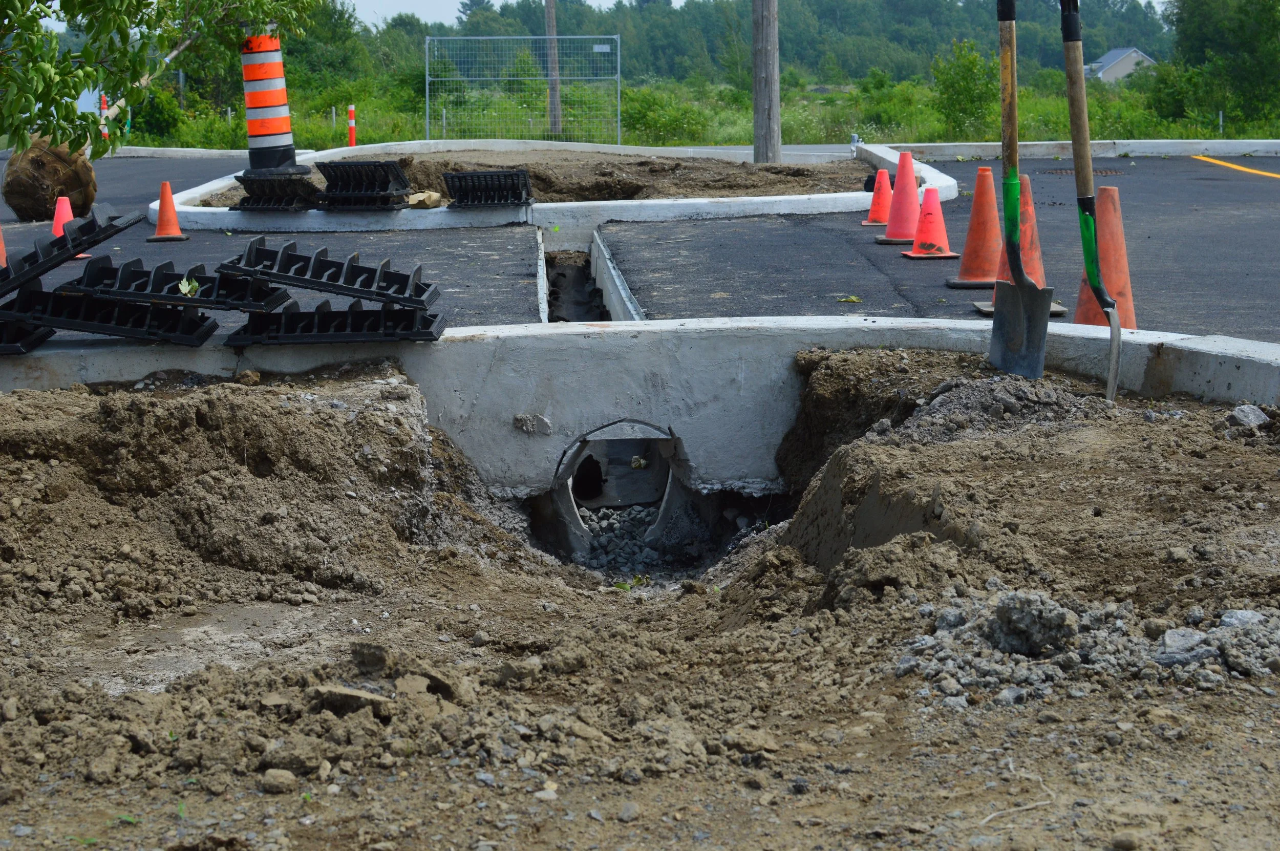 Construction site with dug-up ground, orange cones, black and orange striped cones, and construction tools including shovels and black plastic edging, in a parking lot surrounded by trees and greenery.