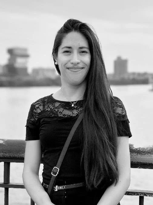 Black and white photo of a young woman with long dark hair, smiling, standing outdoors near a body of water with city buildings in the background.
