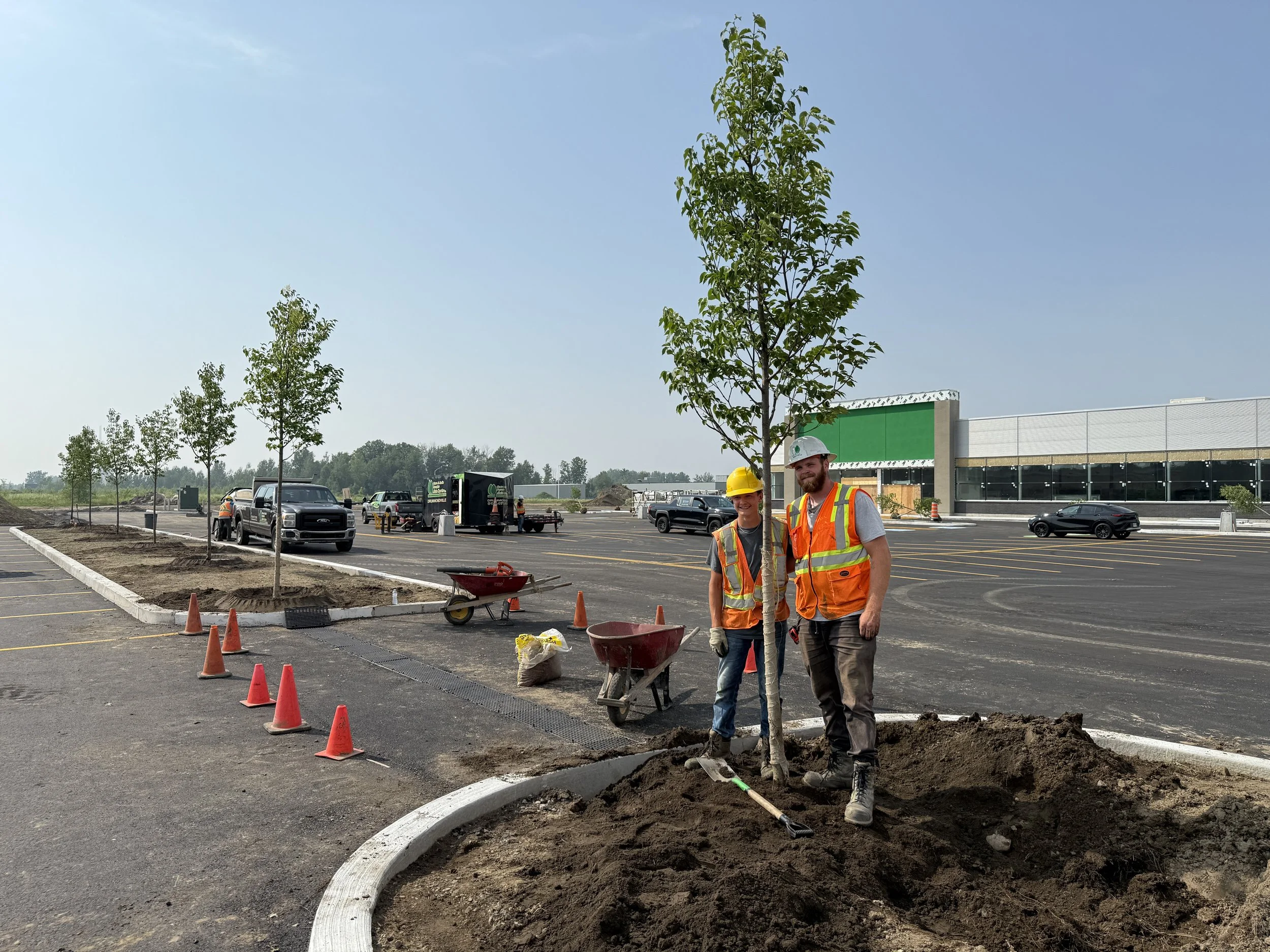 Two construction workers, one woman and one man, wearing safety vests and helmets, standing in a parking lot under construction. They are smiling and pose for the photo with construction tools and equipment around them. There are parked cars, trees, and a building in the background.
