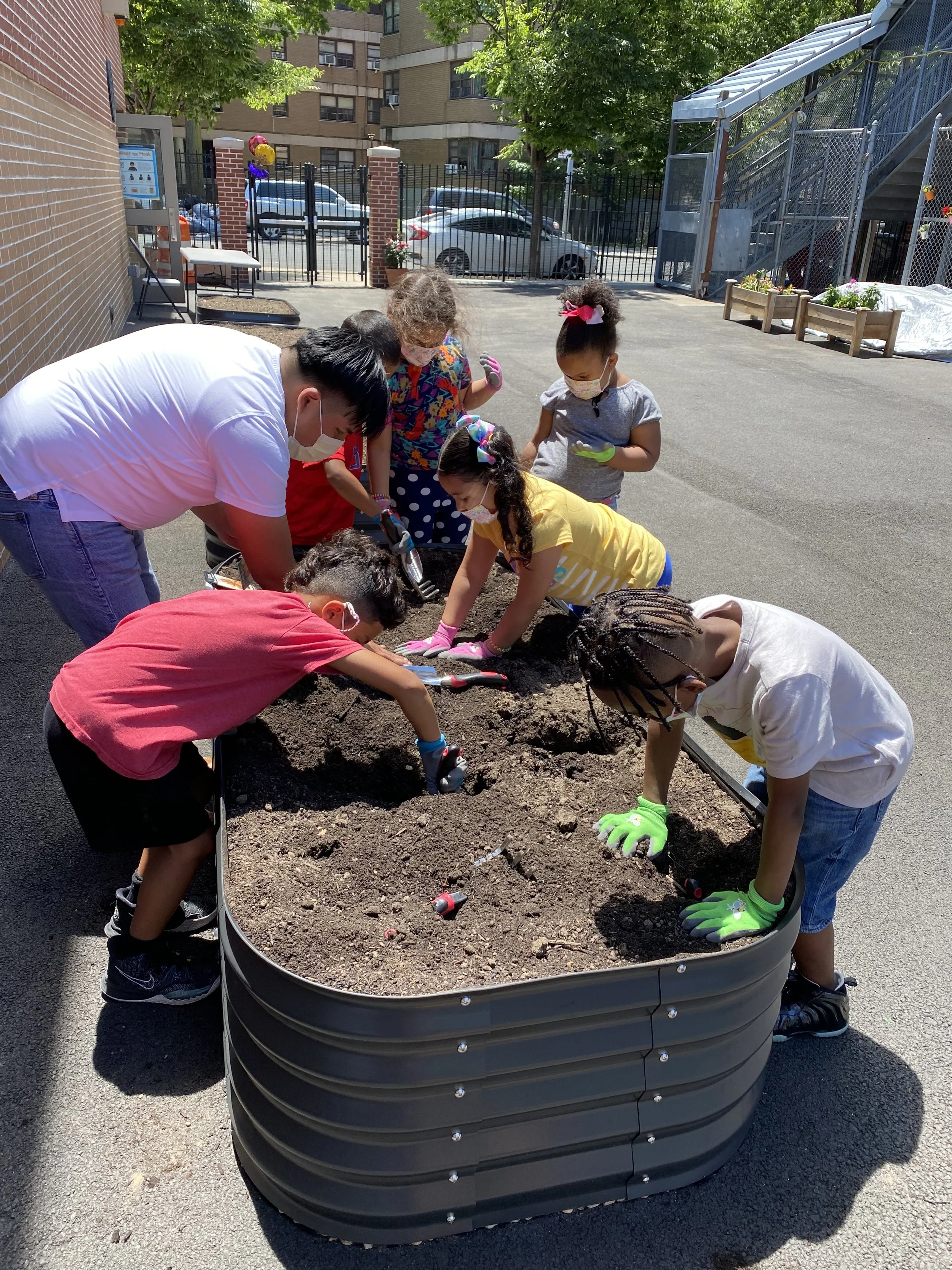 Children and adults planting flowers in a raised garden bed outdoors on a sunny day, with buildings, cars, and trees in the background.