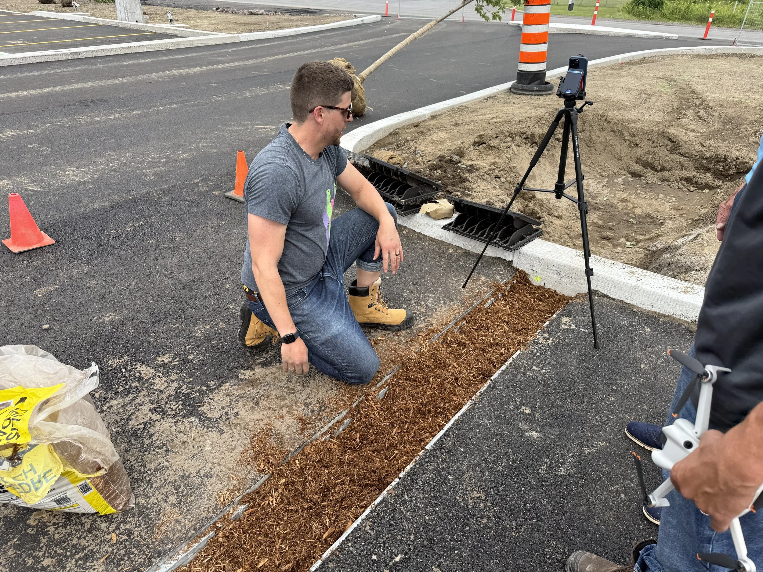 Construction worker kneeling on the ground, inspecting a freshly poured concrete curb with a trench of soil beside it, while a tripod with a measuring device is set up nearby during daytime outdoor construction.