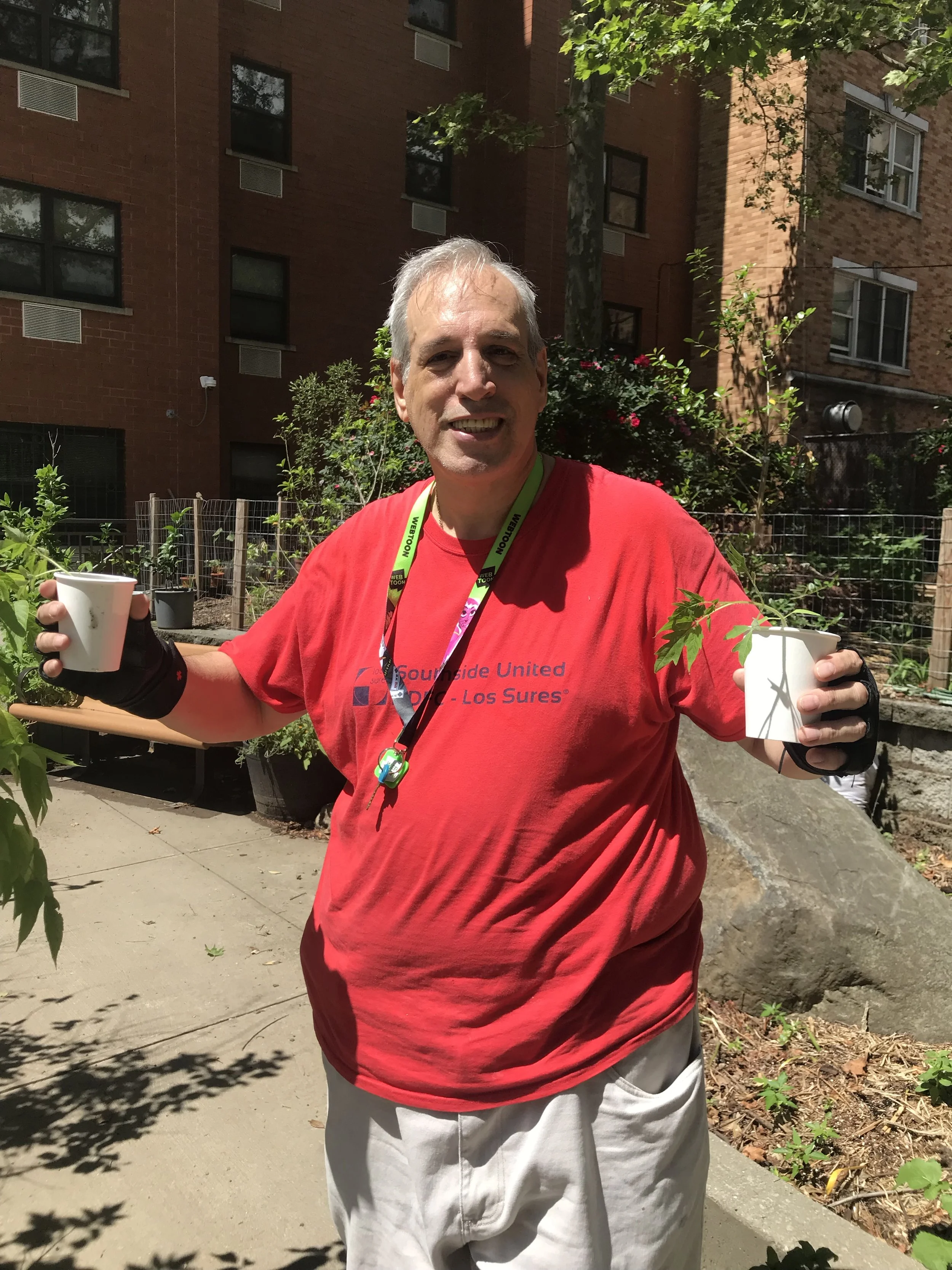 A smiling middle-aged man with gray hair standing outdoors in front of a brick building, holding two white paper cups, wearing a red T-shirt and gray shorts, with trees and shrubs around.
