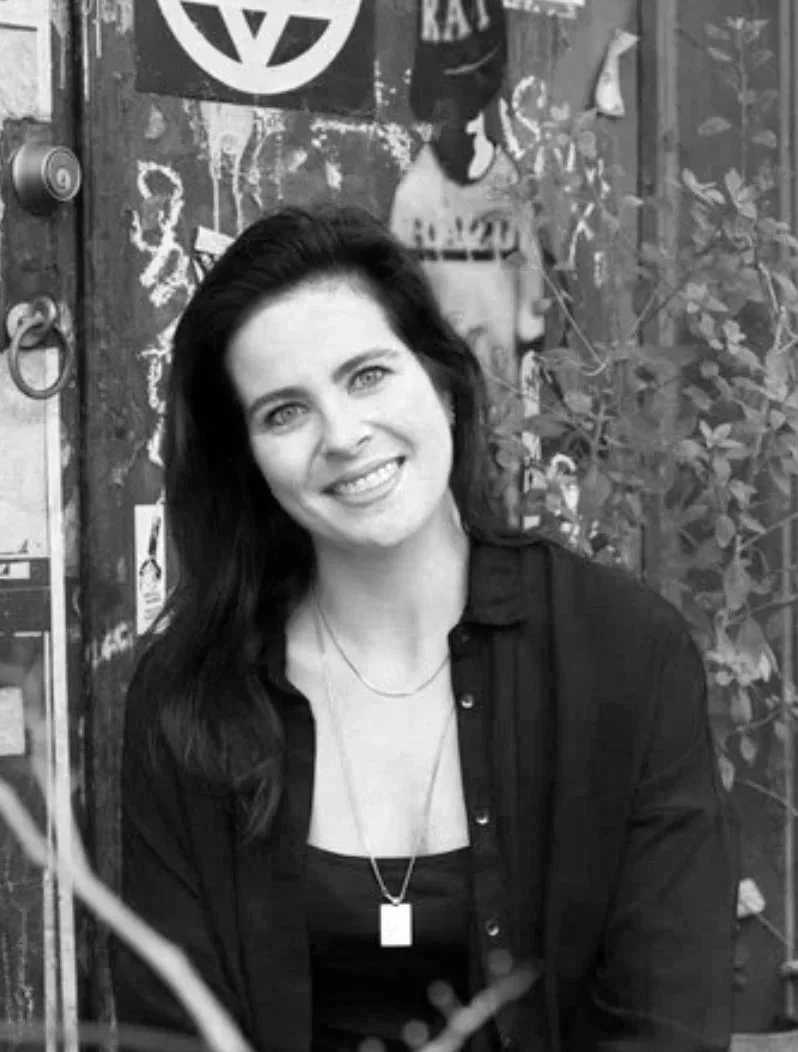 Black and white photo of a woman with dark hair, smiling, wearing a black top and a necklace, sitting in front of a decorated wall with posters and plants.