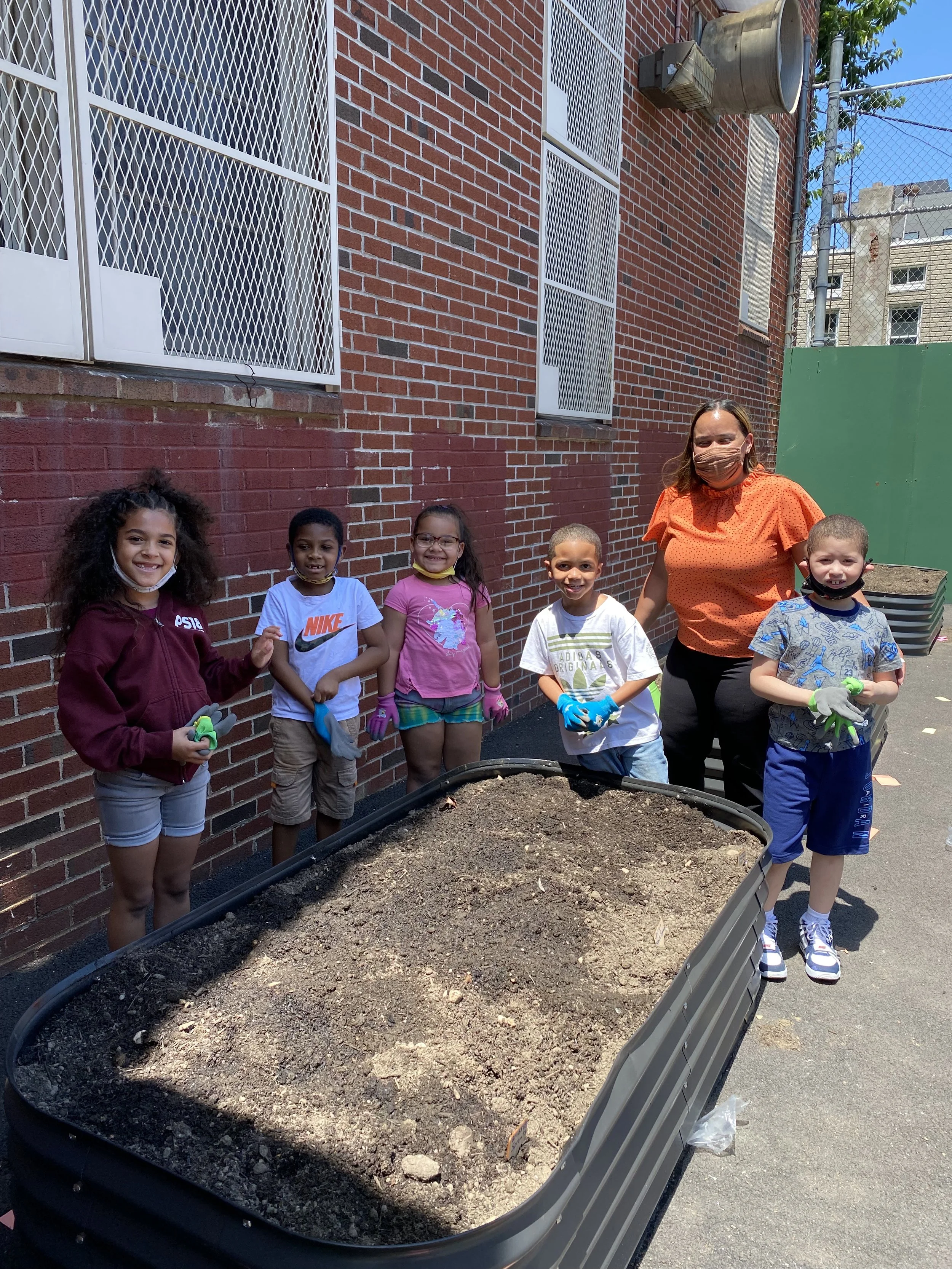 Group of children and a woman standing around a large planter filled with soil outside a brick building, some children wearing gloves.