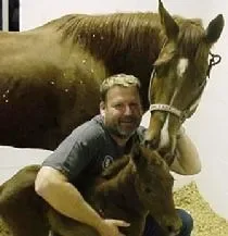 A man smiling and hugging two horses in an indoor setting.