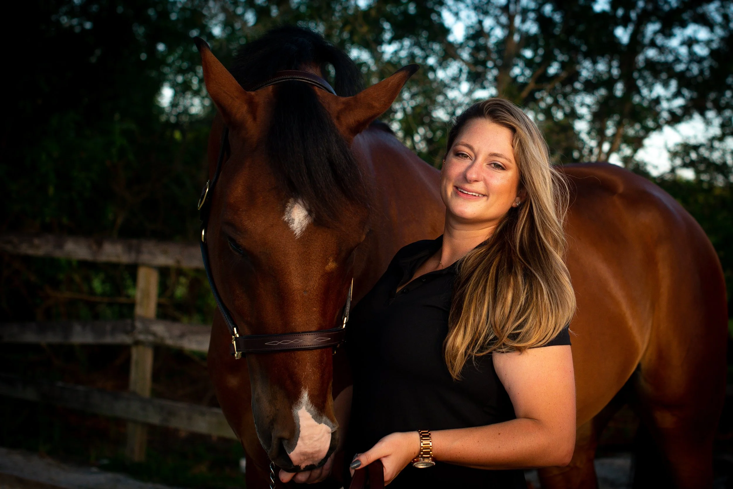A woman with long blonde hair smiling while standing next to and holding a brown horse outside during daytime, with trees and a wooden fence in the background.