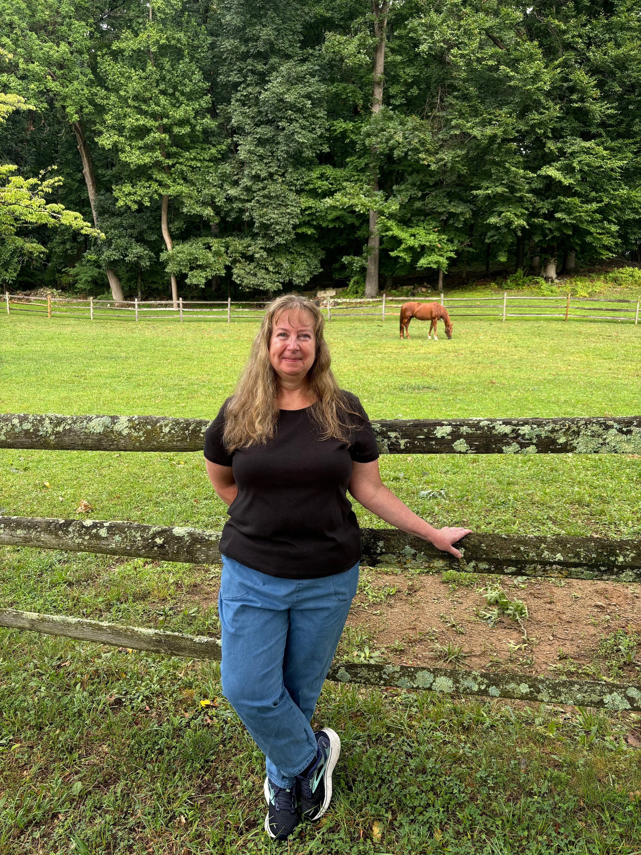 A woman with long, wavy hair standing outdoors in front of a wooden fence, green grass, and trees. She is wearing a black t-shirt, blue jeans, and sneakers, and is leaning against the fence with one hand. There is a brown horse grazing in the background.