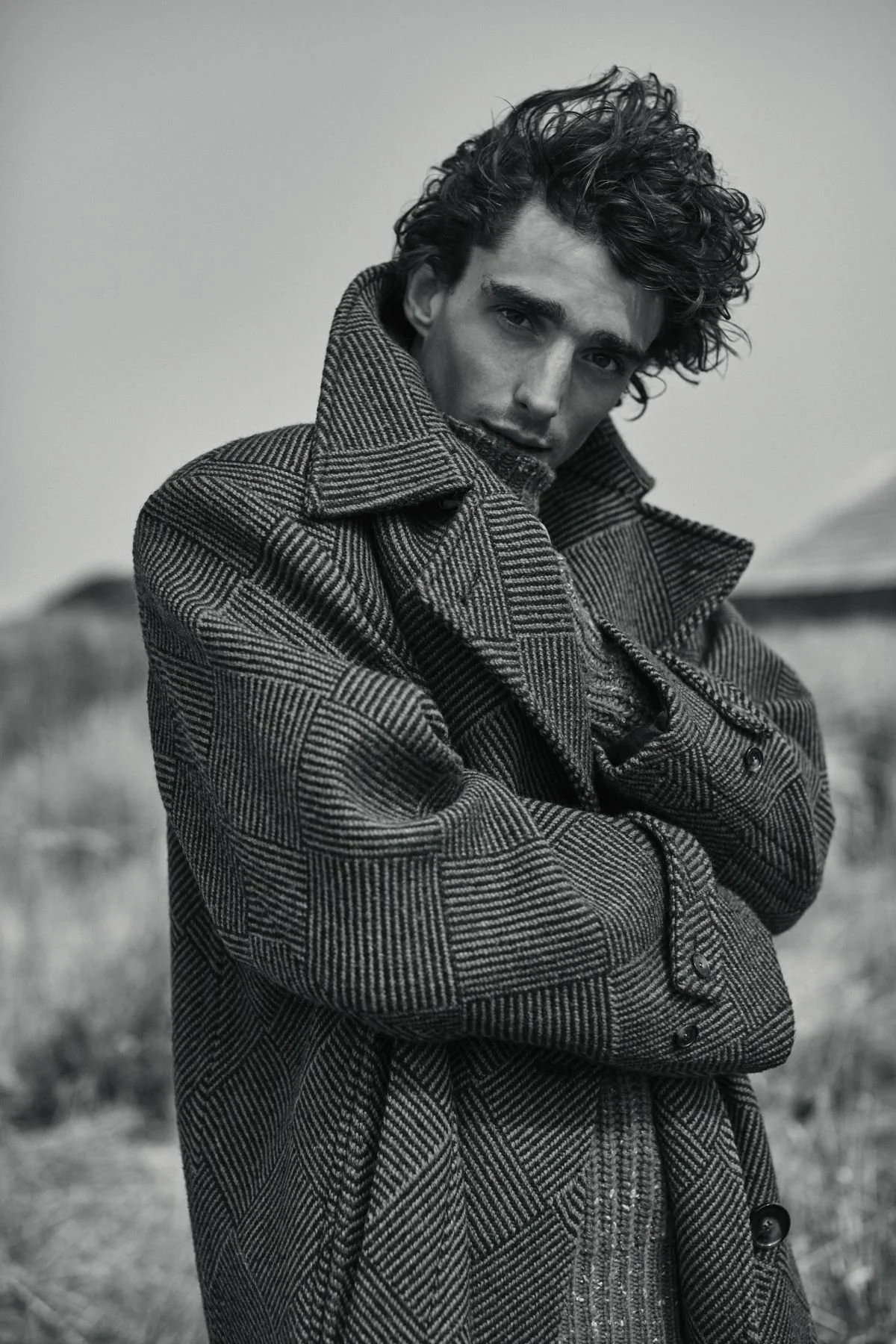A black and white photo of a young man with curly hair wearing a patterned coat outdoors.