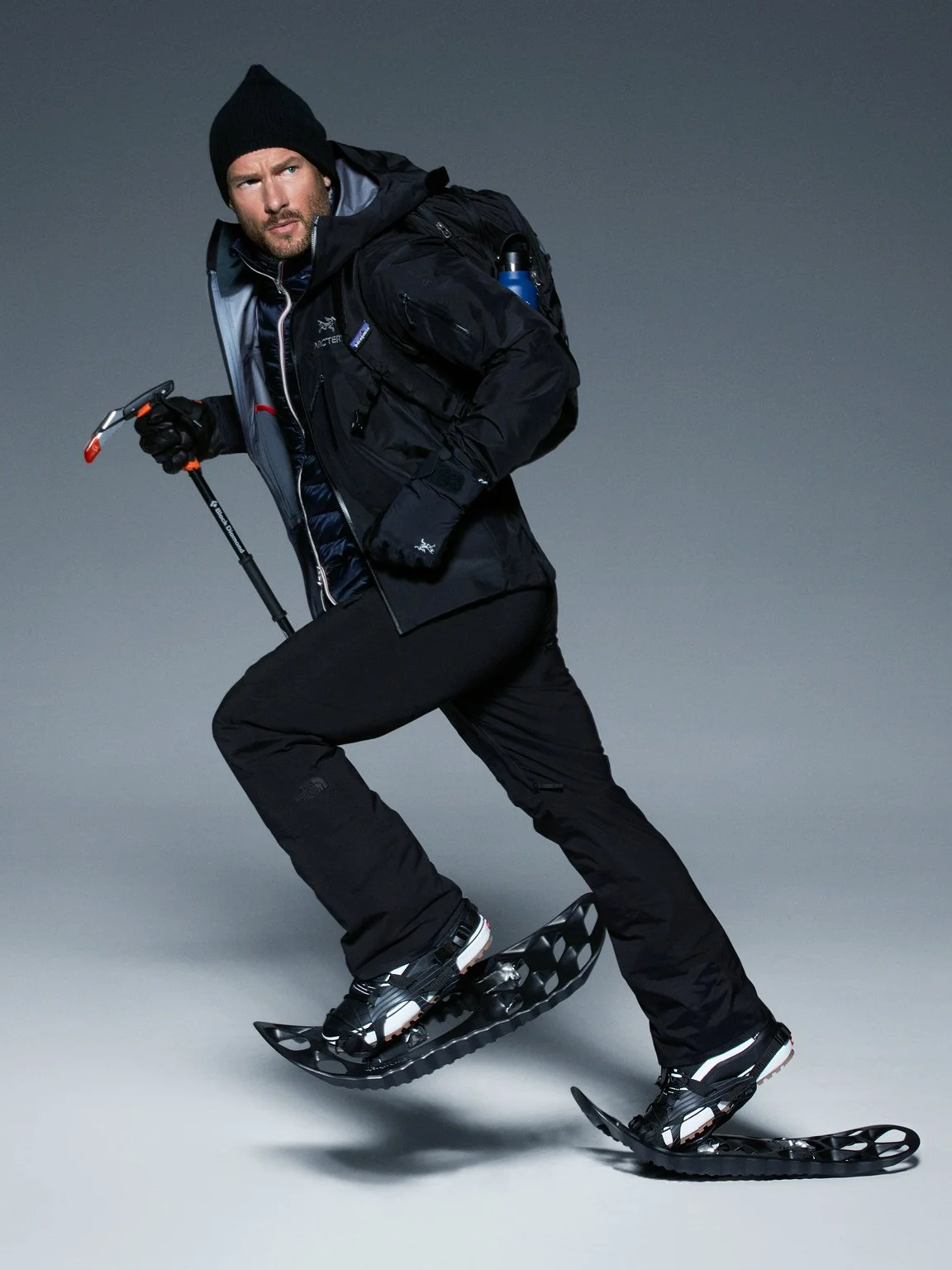 A man dressed in black winter gear snowshoes on a plain gray background.