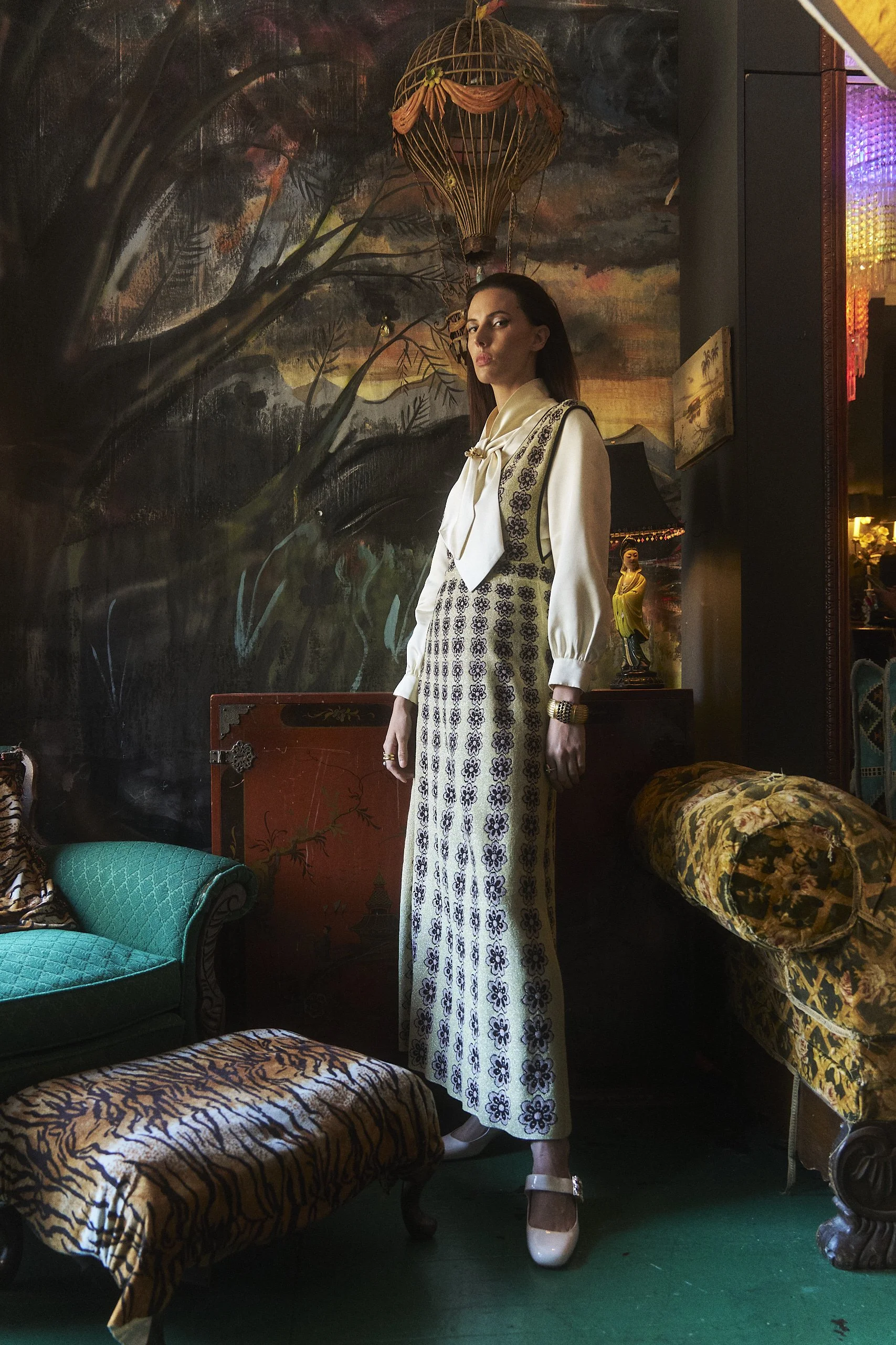 A woman with dark hair wearing a cream-colored blouse and patterned high-waisted pants, standing indoors in front of a dark, painted wall with a landscape scene. The room has eclectic decor including a hot air balloon wall hanging, a statue of a woma
