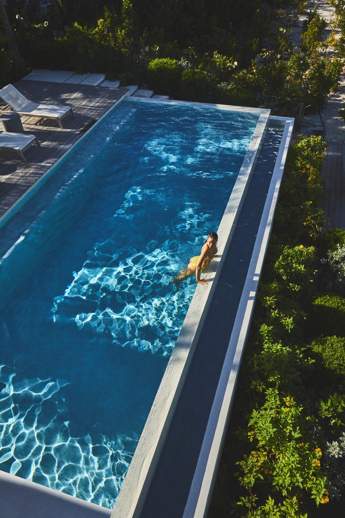 A woman in a yellow swimsuit relaxing in an outdoor swimming pool with clear blue water, surrounded by greenery and lounge chairs.