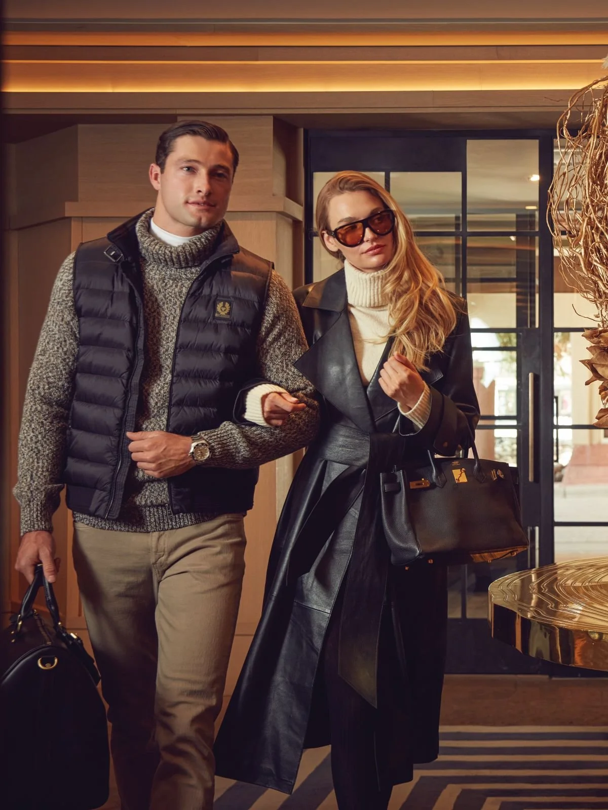 A man and woman walking into a hotel or lobby. The man is wearing a black puffer vest over a gray sweater, beige pants, and a watch. The woman is wearing a black leather jacket, black wide-leg pants, large sunglasses, and carrying a large black handb