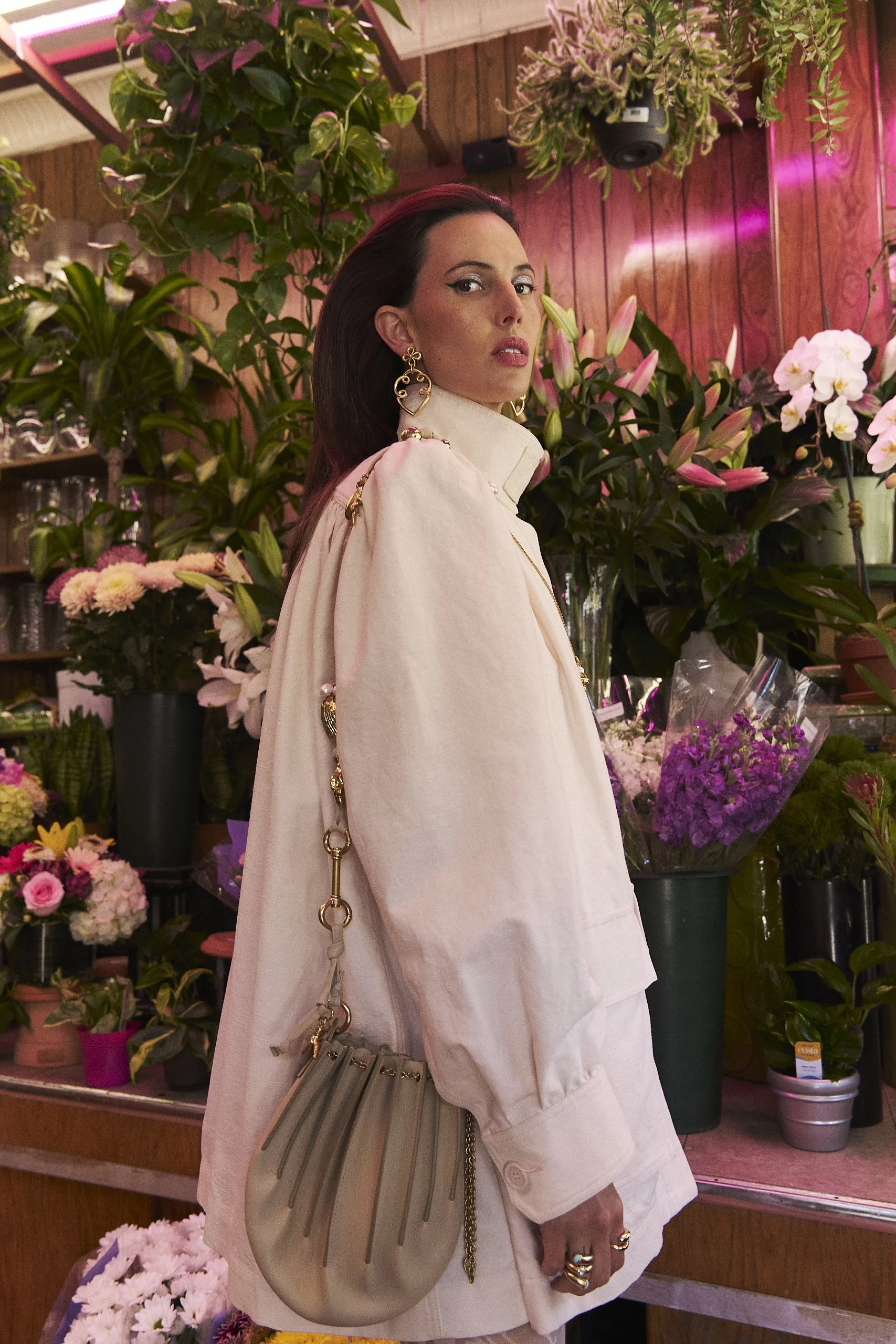 A woman with dark hair, wearing statement earrings and a light-colored oversized blazer, stands in a flower shop surrounded by various colorful flowers and green plants.