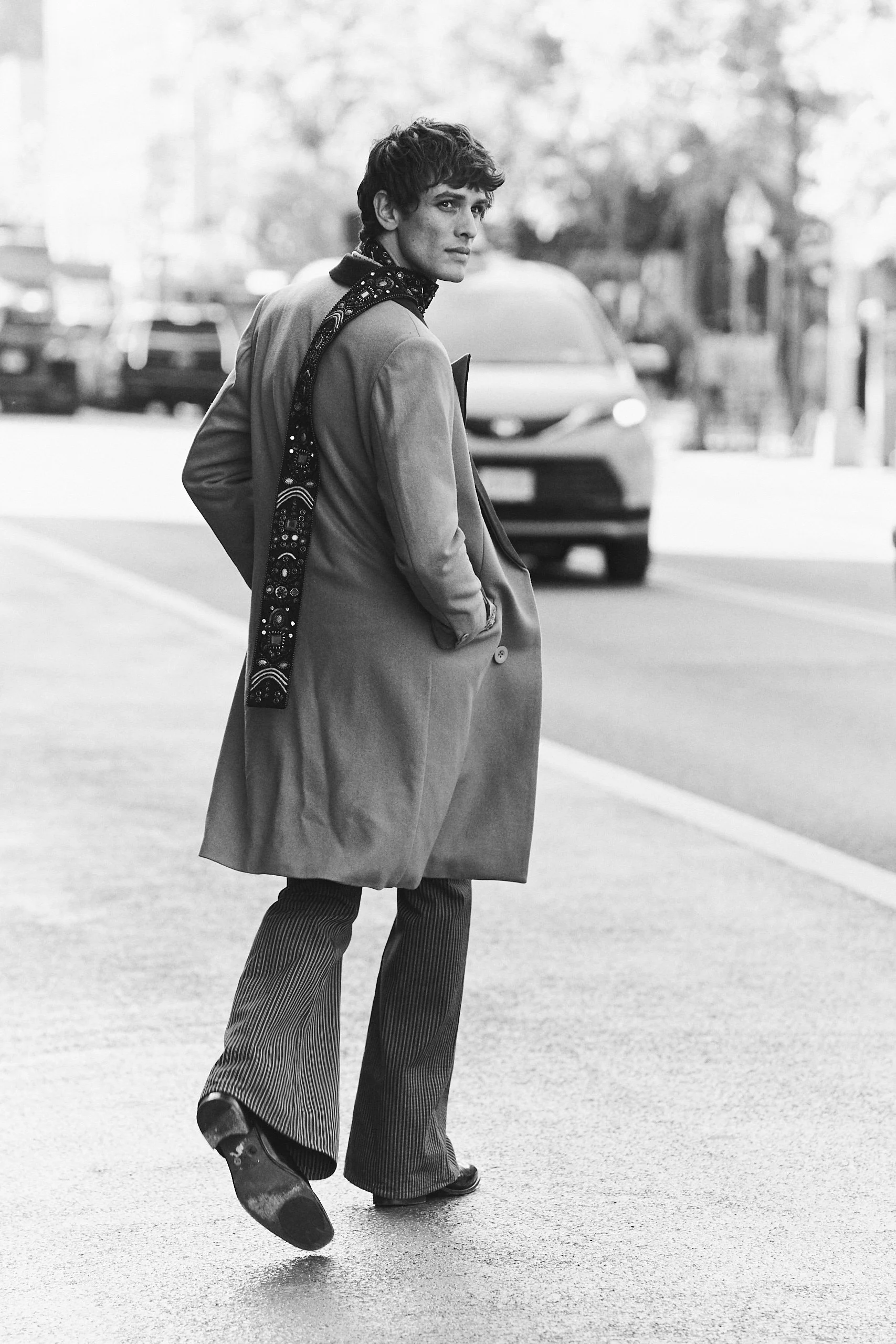 Black and white photo of a young man with dark, curly hair looking over his shoulder while walking on a street with cars in the background. He is wearing a long coat, patterned pants, and a bandana around his neck.