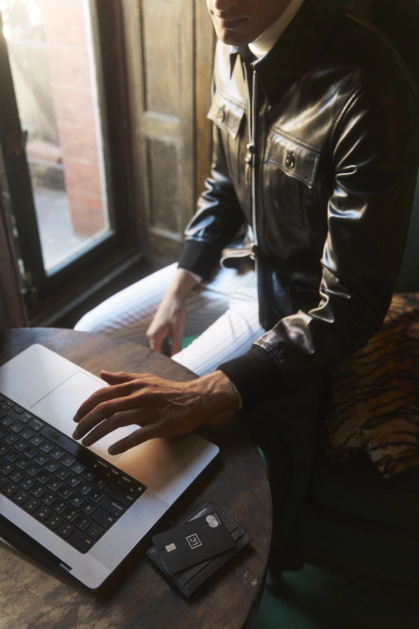 Close-up of a person in a black leather jacket working on a laptop at a wooden table, with a credit card and a black wallet nearby, near a window with natural light.
