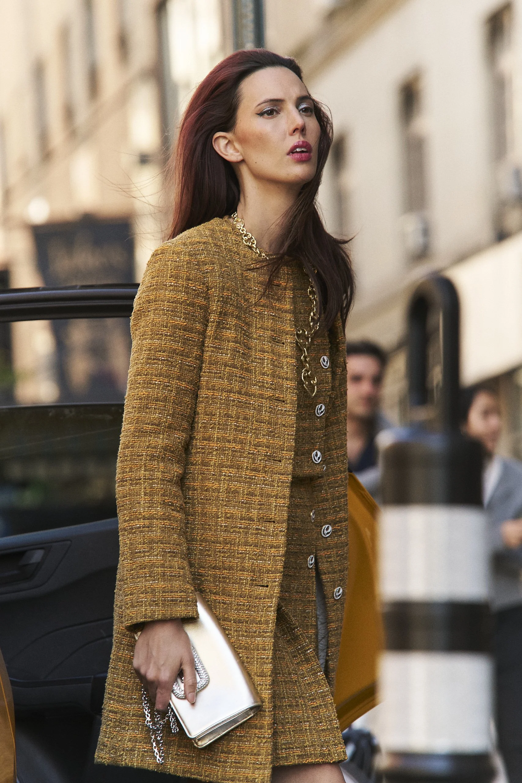 A woman with long dark hair wearing a golden-brown textured coat with buttons, holding a white clutch purse with a chain strap, standing outdoors with a blurred city background.