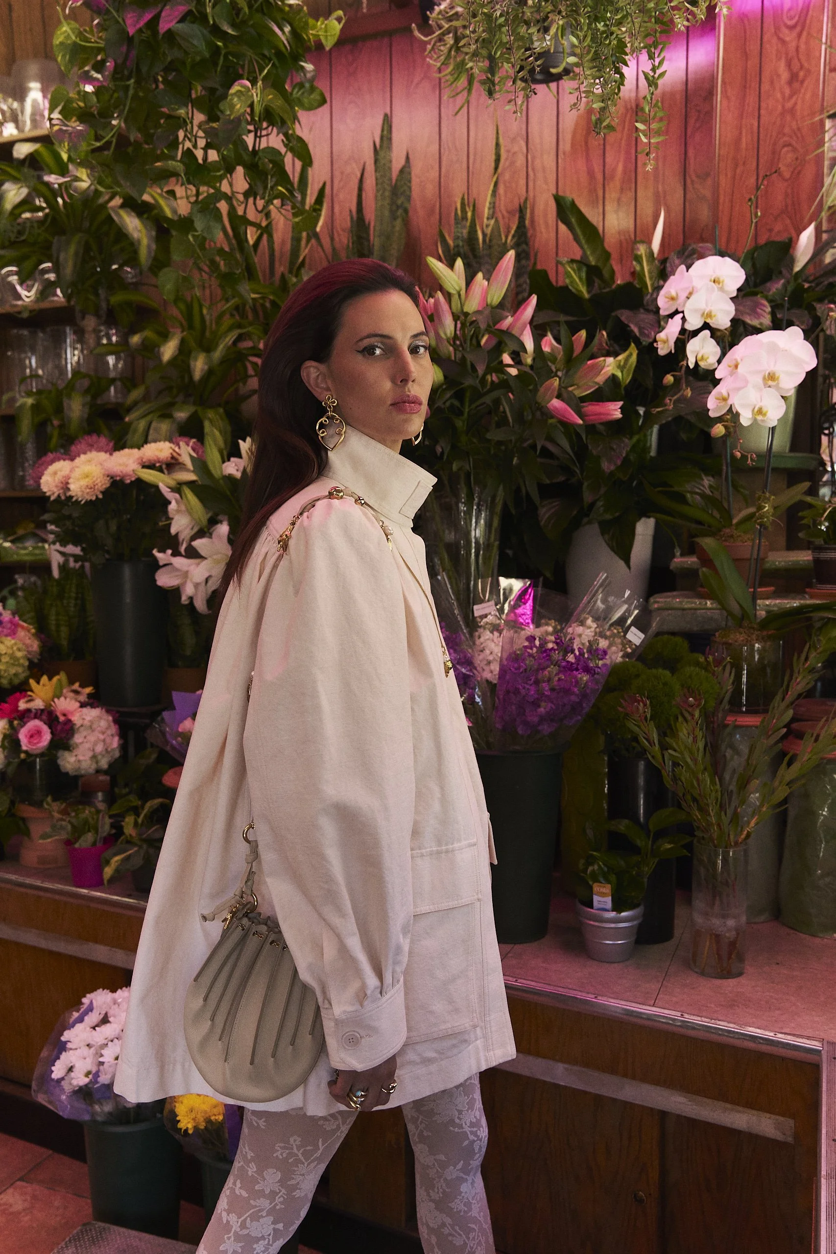 A woman with dark red hair dressed in beige, standing in a flower shop surrounded by various pink and white flowers and green plants.
