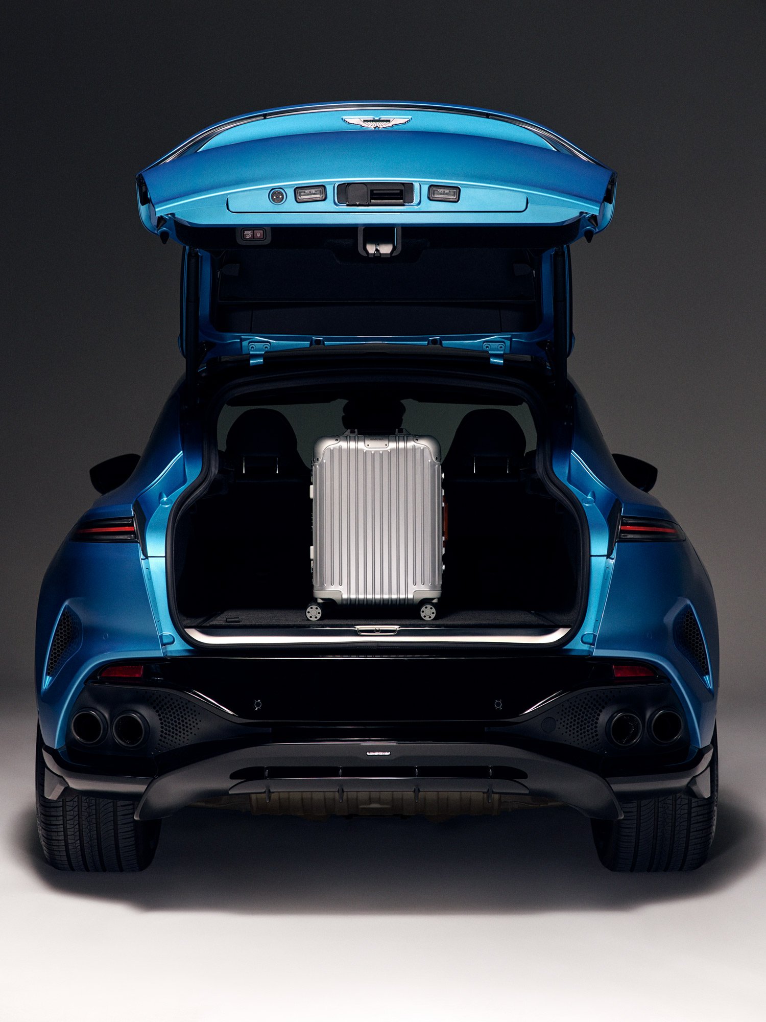 The rear of a blue electric vehicle with its trunk open, revealing a silver suitcase inside. The car is set against a dark background.