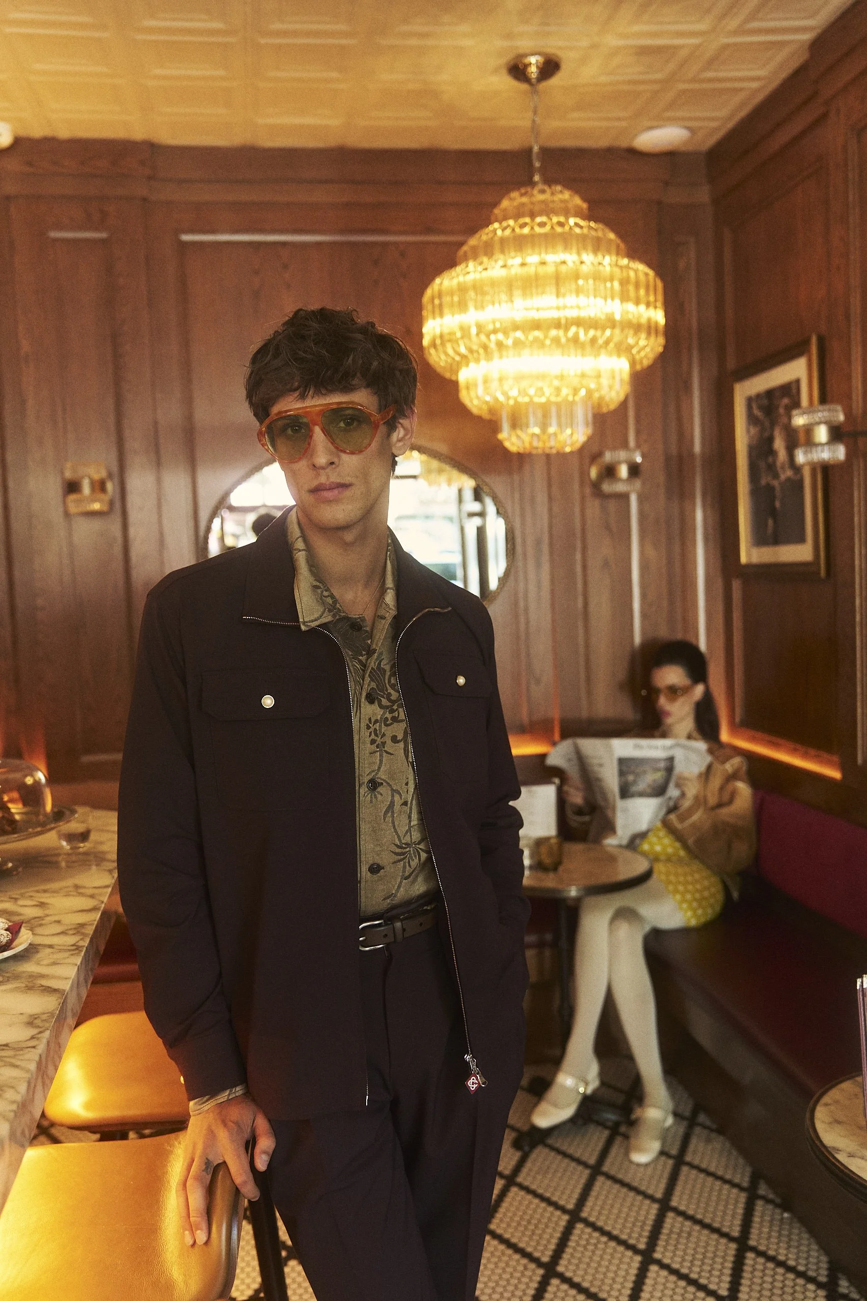 A young man with brown curly hair wearing orange sunglasses, a patterned shirt, and a black jacket standing in a vintage-style restaurant or cafe with wood-paneled walls and a large chandelier. In the background, a woman sitting at a table is reading