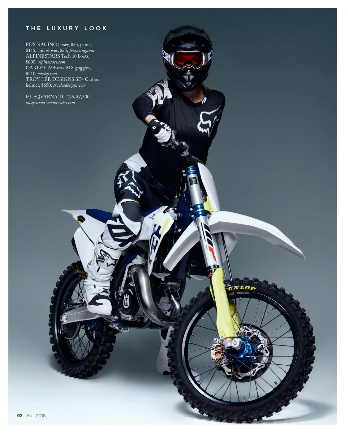 A person dressed in motocross gear riding a Husqvarna dirt bike, featuring a black helmet with red goggles, black and white Fox Racing apparel, and a white motocross bike with yellow and blue accents.