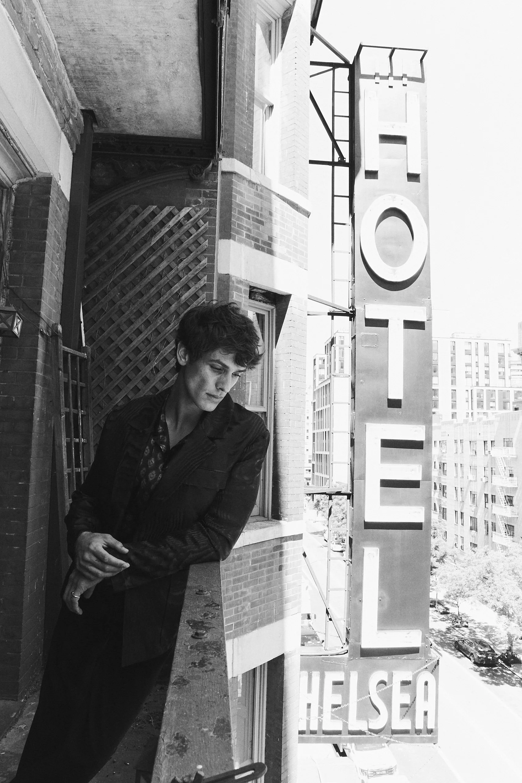 A young man with curly hair in a dark jacket standing on a city balcony next to a large vertical sign that reads 'HELSEA' in capital letters. Buildings and a street with parked cars are visible in the background.