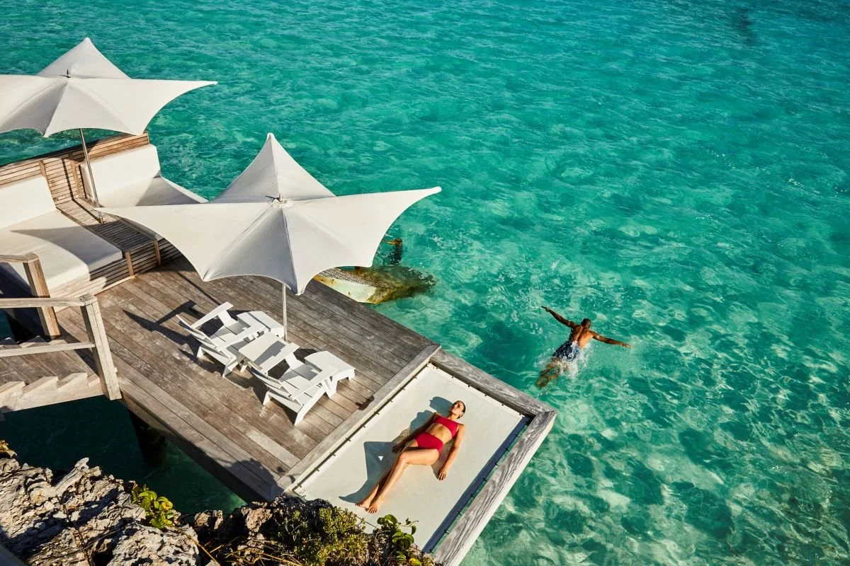 A woman in a red swimsuit is lying on a lounge chair on a wooden deck overlooking turquoise water. There are two large white umbrellas providing shade. A man is swimming in the water nearby.
