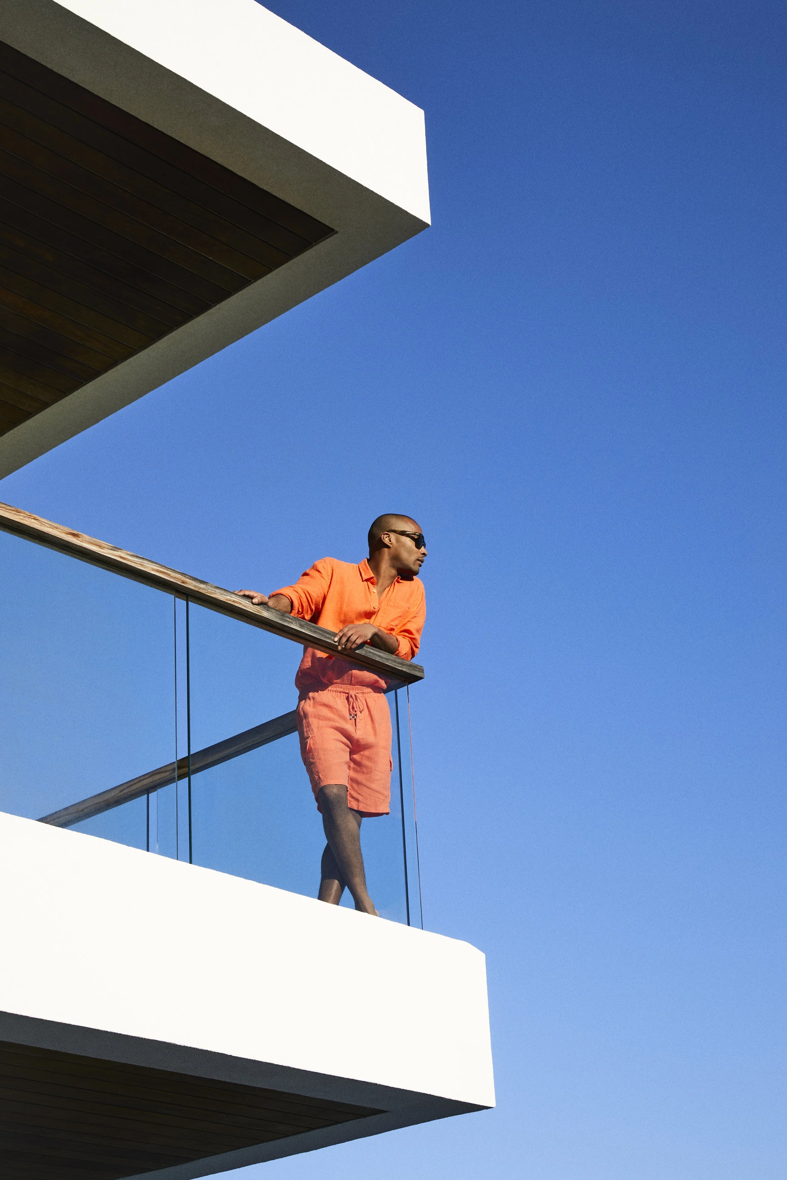 A man in an orange shirt and shorts stands on a balcony with glass railings, looking to the right, against a clear blue sky.