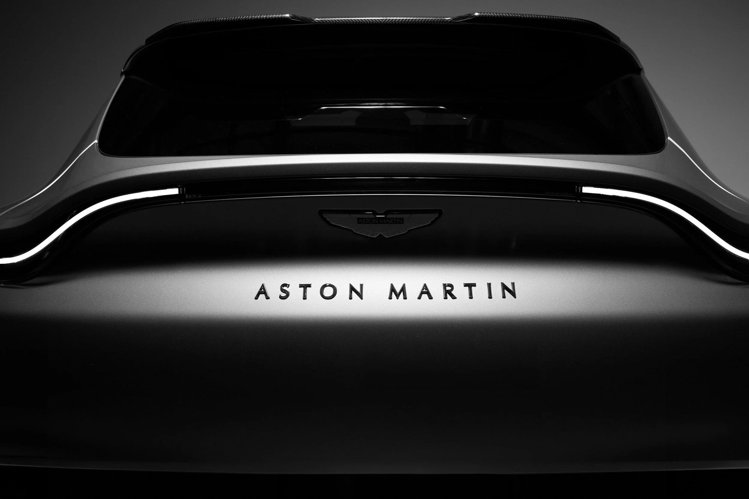 Close-up of the rear of a black Aston Martin car with illuminated taillights and the Aston Martin logo, in a dark environment.