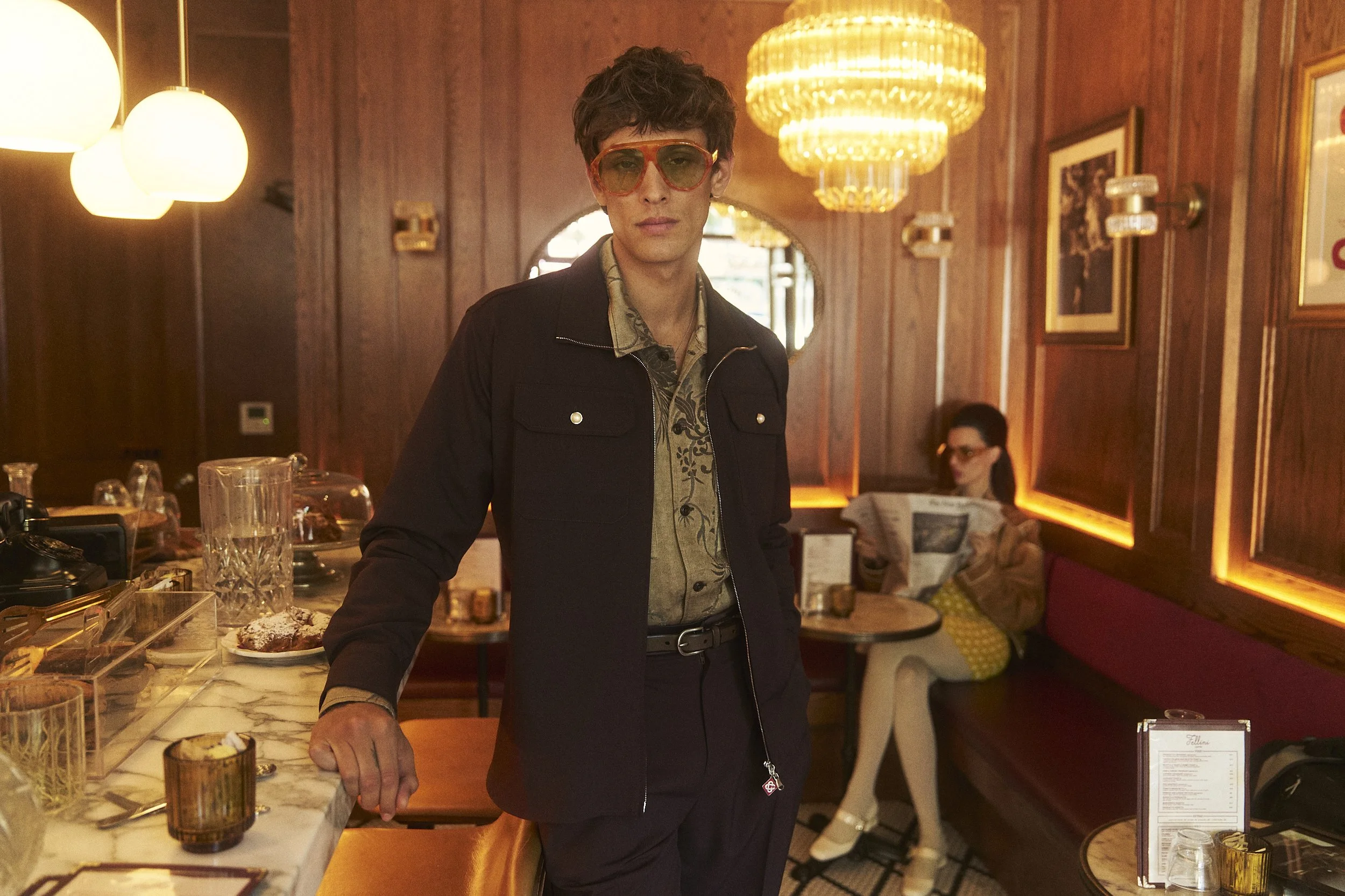 A young man with dark, curly hair and large orange sunglasses stands in a warmly lit restaurant, wearing a black jacket over a patterned shirt. He is positioned at a marble-topped bar or counter with various glassware and desserts nearby. The backgro