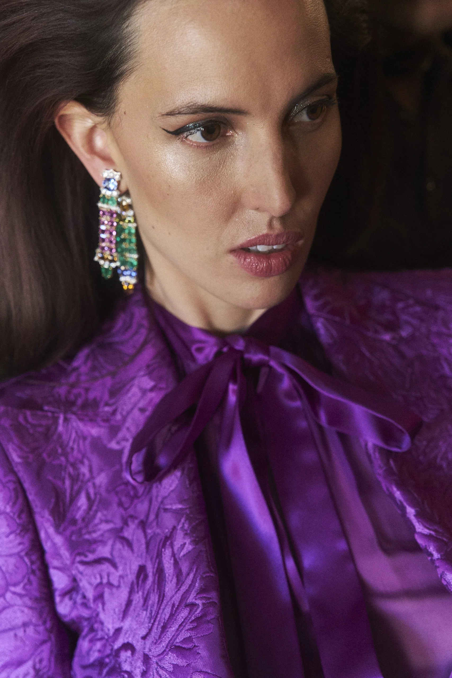 A close-up of a woman with long brown hair, wearing colorful dangling earrings, a purple patterned blouse with a bow at the neckline, and makeup with winged eyeliner and light lipstick.