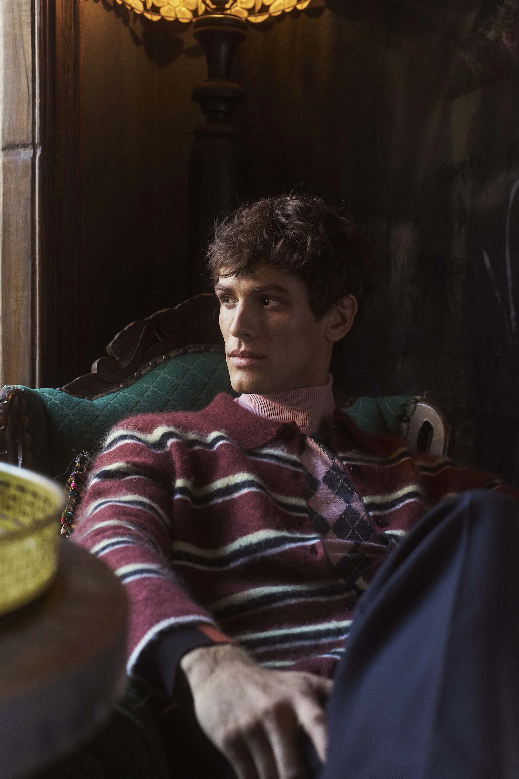 A young man with curly brown hair sitting on a green and brown patterned chair, wearing a red and white striped sweater and a pink turtleneck shirt underneath, looking to his left. The room has dark wooden walls and a warm lamp above.