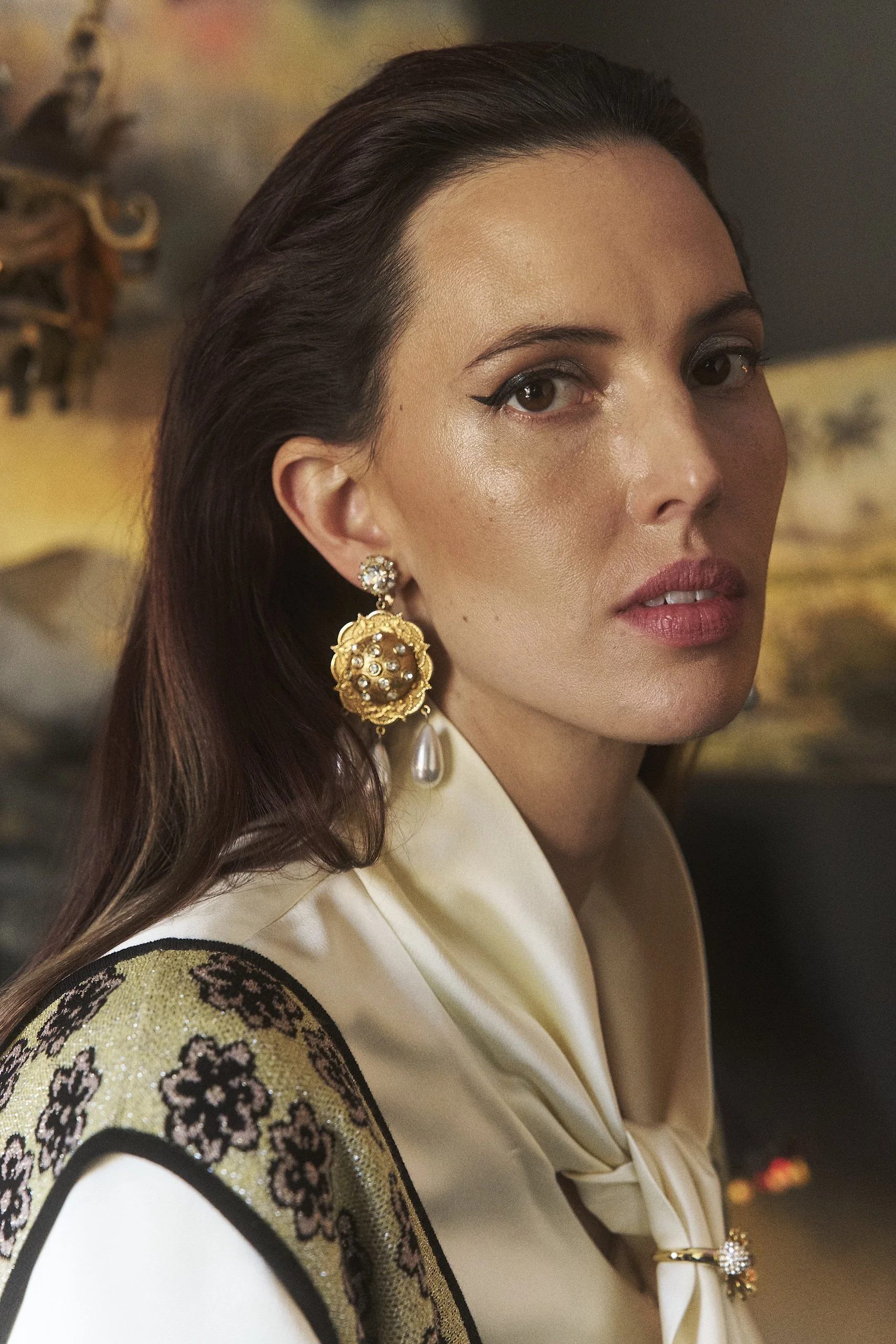 Close-up of a woman with dark hair, wearing large gold and pearl earrings, a cream-colored satin top with a bow, and black eyeliner, against a blurred background.