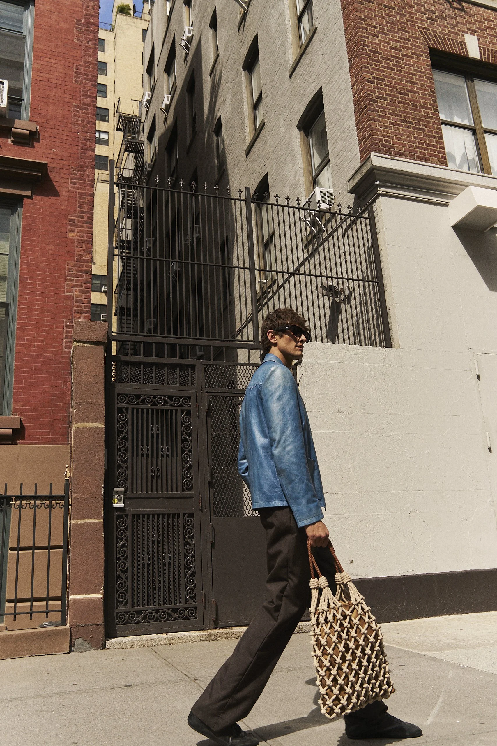 A person wearing a blue jacket, black pants, and sunglasses walking on the sidewalk, carrying a large woven tote bag, with tall city buildings in the background.