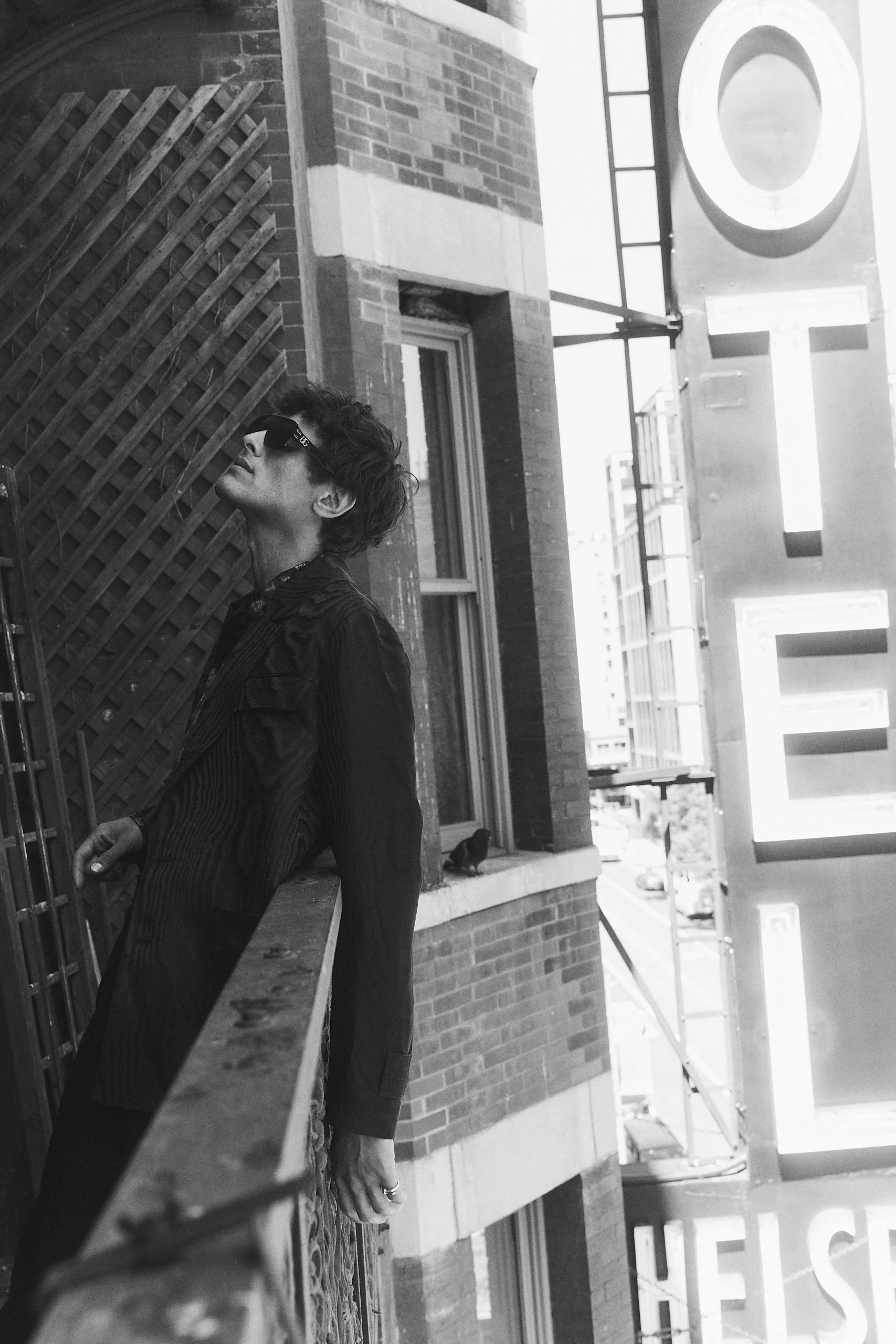 A young man with dark, curly hair and sunglasses leaning against a railing outside a brick building in an urban area.