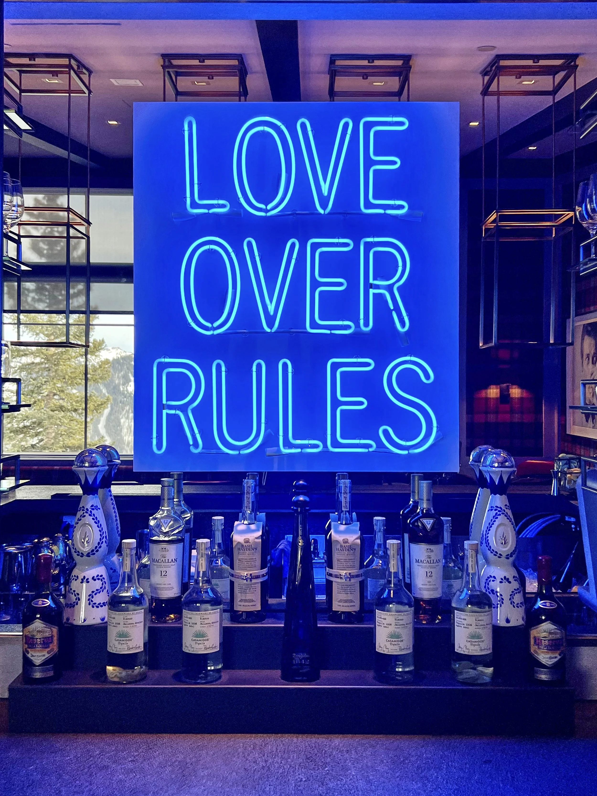 Neon sign reading 'LOVE OVER RULES' in blue, with a bar countertop in front displaying several bottles of alcohol and decorative ceramic bottles.