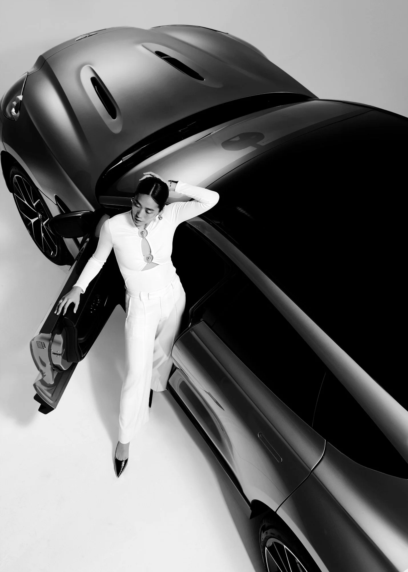 A woman in white outfit and heels stands next to a sleek, black modern car, viewed from above in a studio setting.