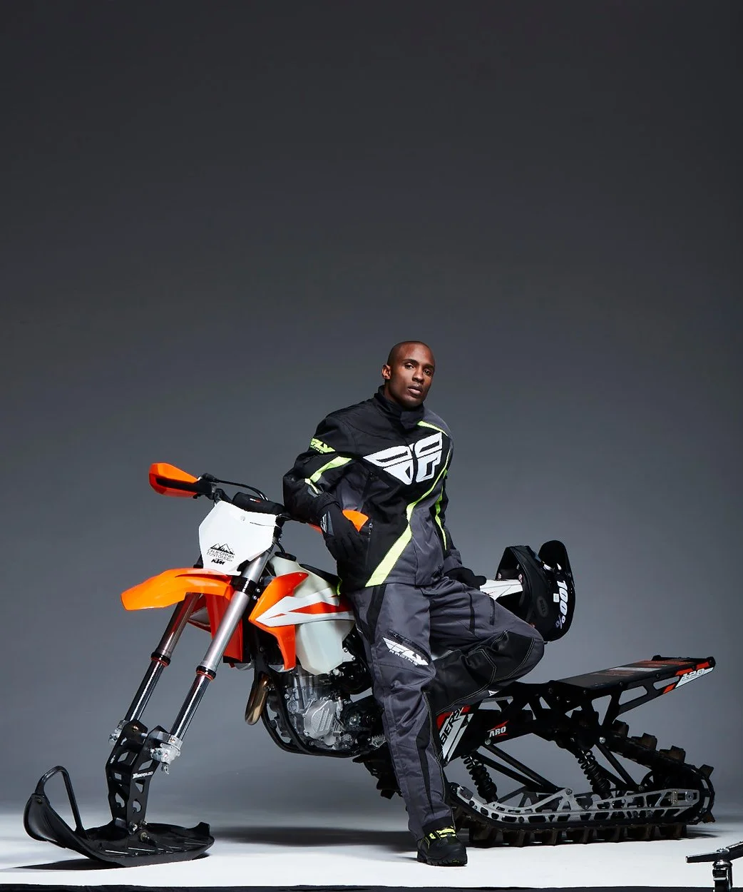 A man in motorcycle gear standing next to an orange and white snow bike with a black helmet hanging on the bike. The background is plain gray.