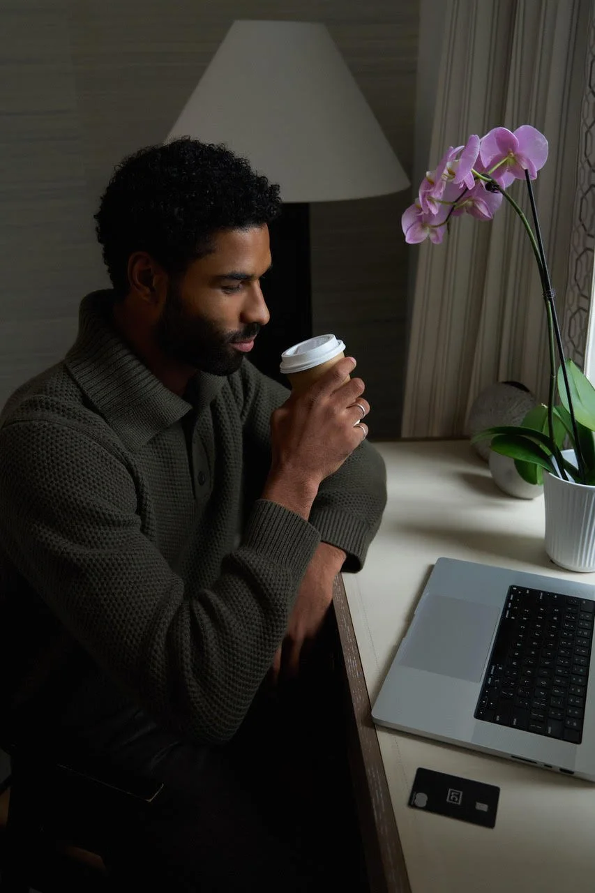 Man sitting at a desk, holding a coffee cup, with a laptop, a potted orchid, and a credit card nearby.