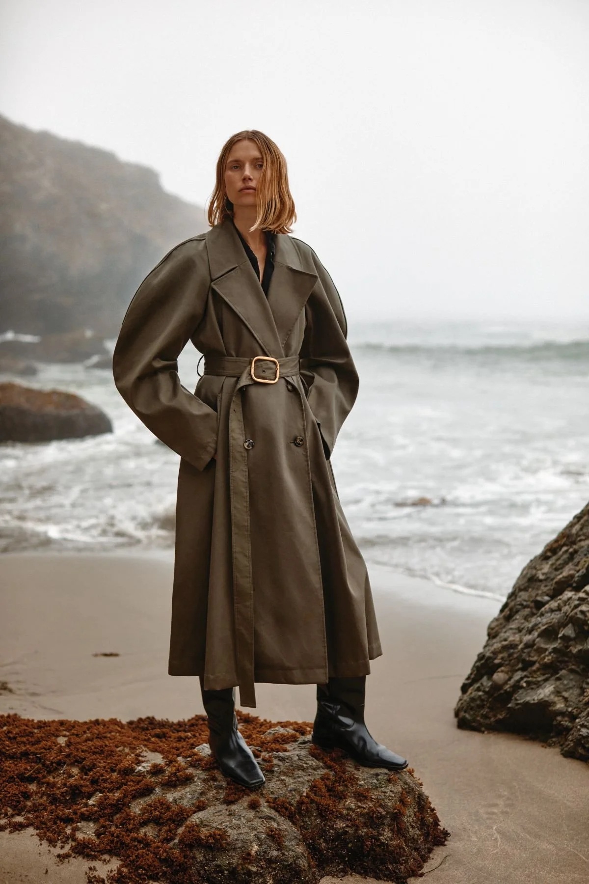 A woman in a long gray trench coat with a belt, standing on a rocky beach with ocean waves and fog in the background.