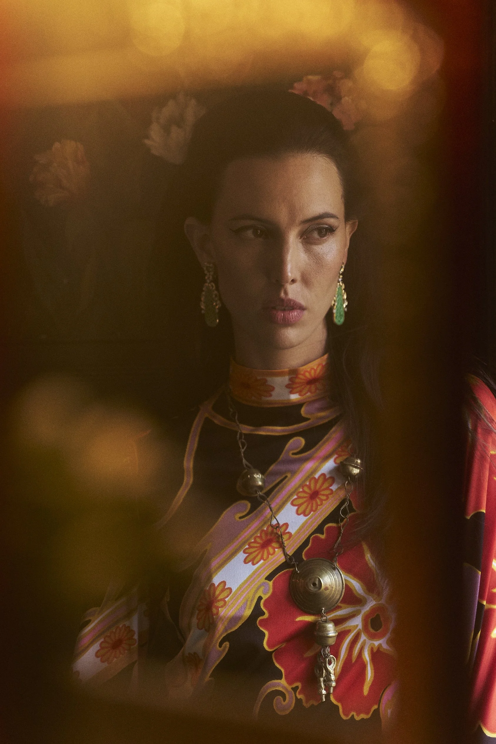 A woman with dark hair styled back, wearing elaborate earrings, a patterned dress with floral motifs, and a necklace with large metallic bells. The background features warm, soft lighting with a golden glow.