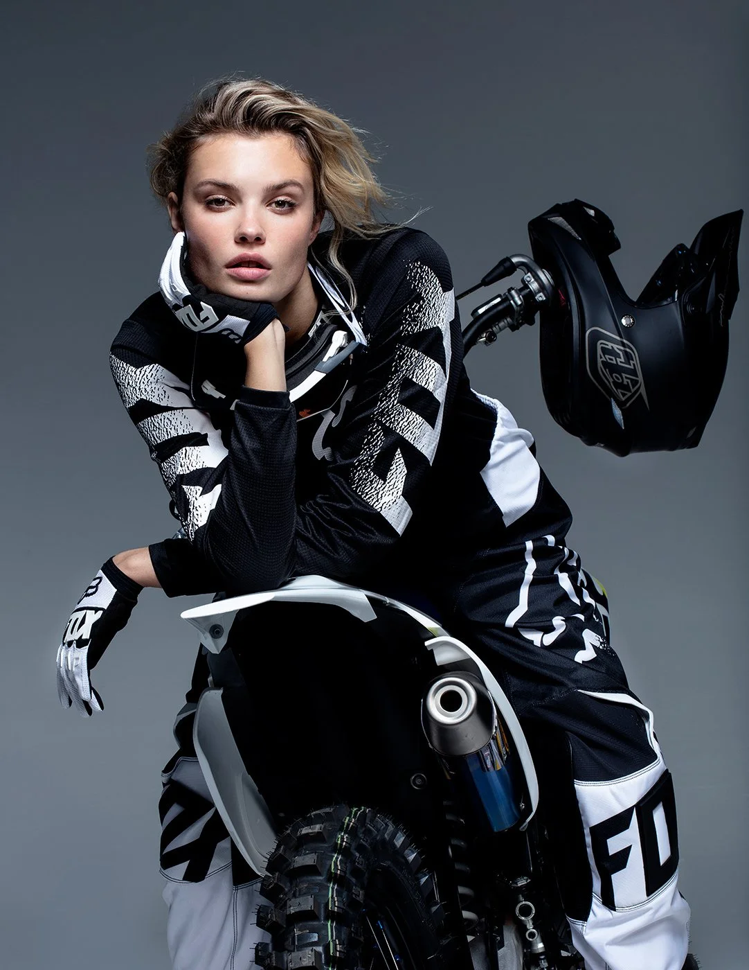 A woman wearing motocross gear, resting her chin on her hand, next to a dirt bike and helmet.