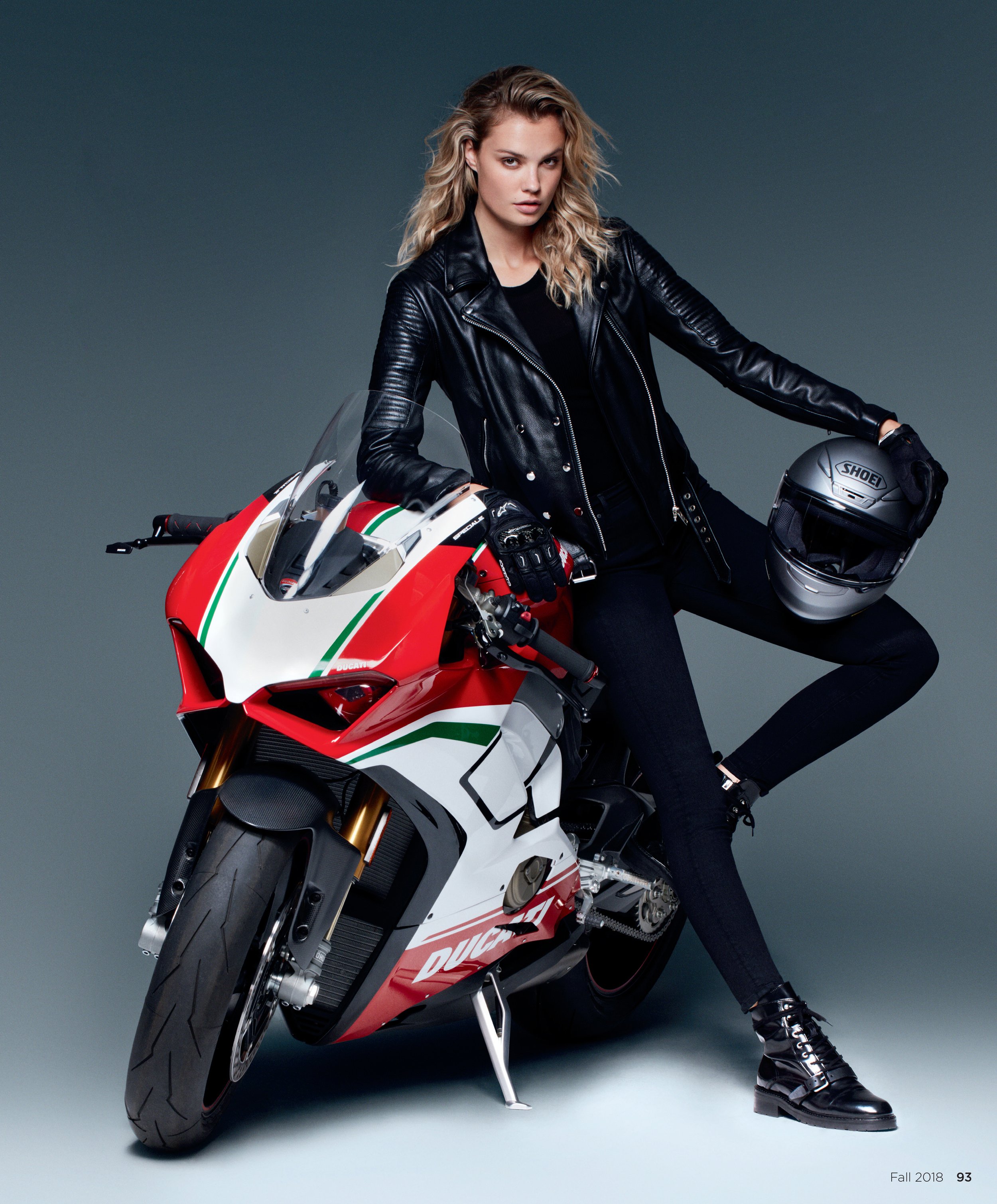 A woman with blonde wavy hair, dressed in black leather jacket, black pants, and black combat boots, sitting on a red and white Ducati motorcycle with a full face helmet in her right hand, against a plain dark gray background.