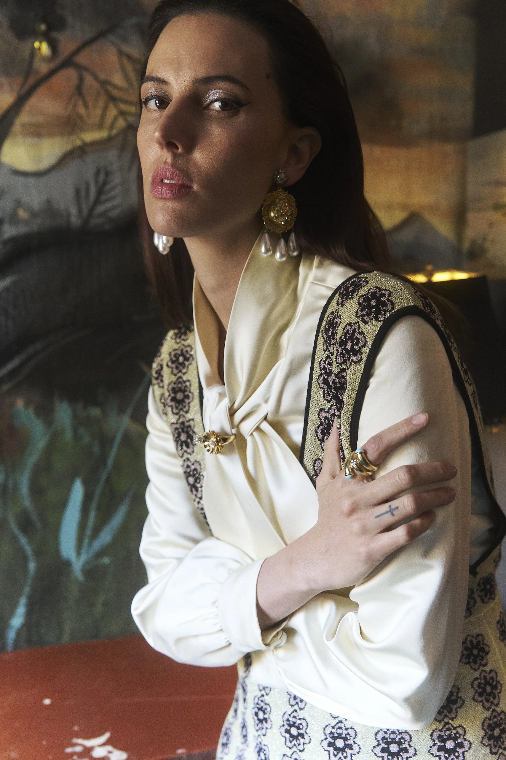 A woman with dark hair, wearing gold and pearl earrings and a cream-colored satin blouse with embroidered floral patterns, stands with her arms crossed in front of a painted wall featuring floral and landscape motifs.