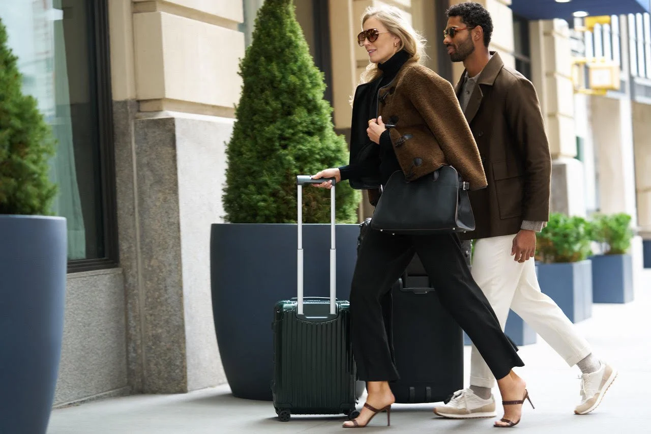 A woman and a man walking on a city sidewalk with rolling suitcases. The woman wears sunglasses, a brown coat, black pants, and high heels, while the man wears glasses, a brown coat, and white pants.