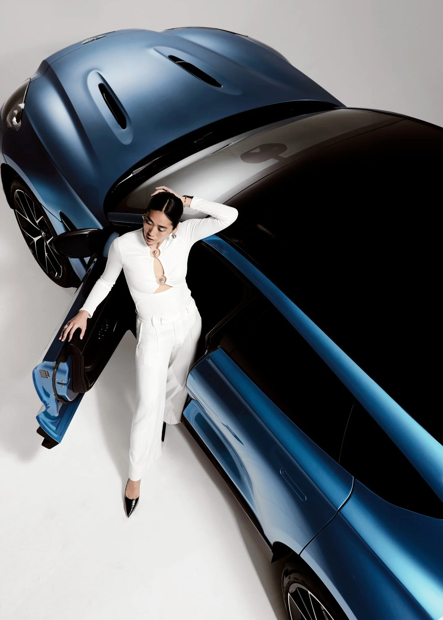A woman in white pants and black heels standing next to a metallic blue sports car, with the car door open, in a bright studio setting.