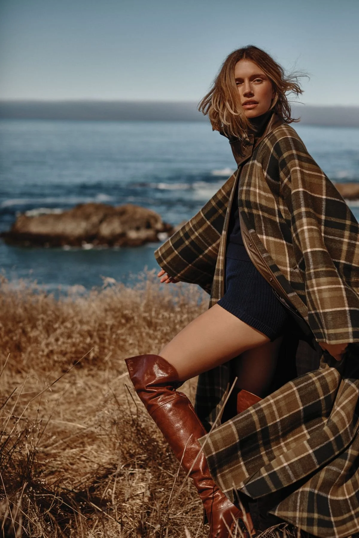 A woman with shoulder-length brown hair wearing a plaid coat, black top, navy shorts, and brown boots, sitting outdoors on dry grass near the ocean under a clear sky.