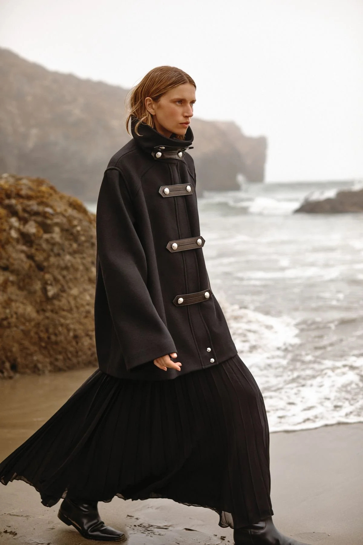 A woman in a black coat and long black pleated skirt walking along a beach with cliffs in the background during foggy weather.