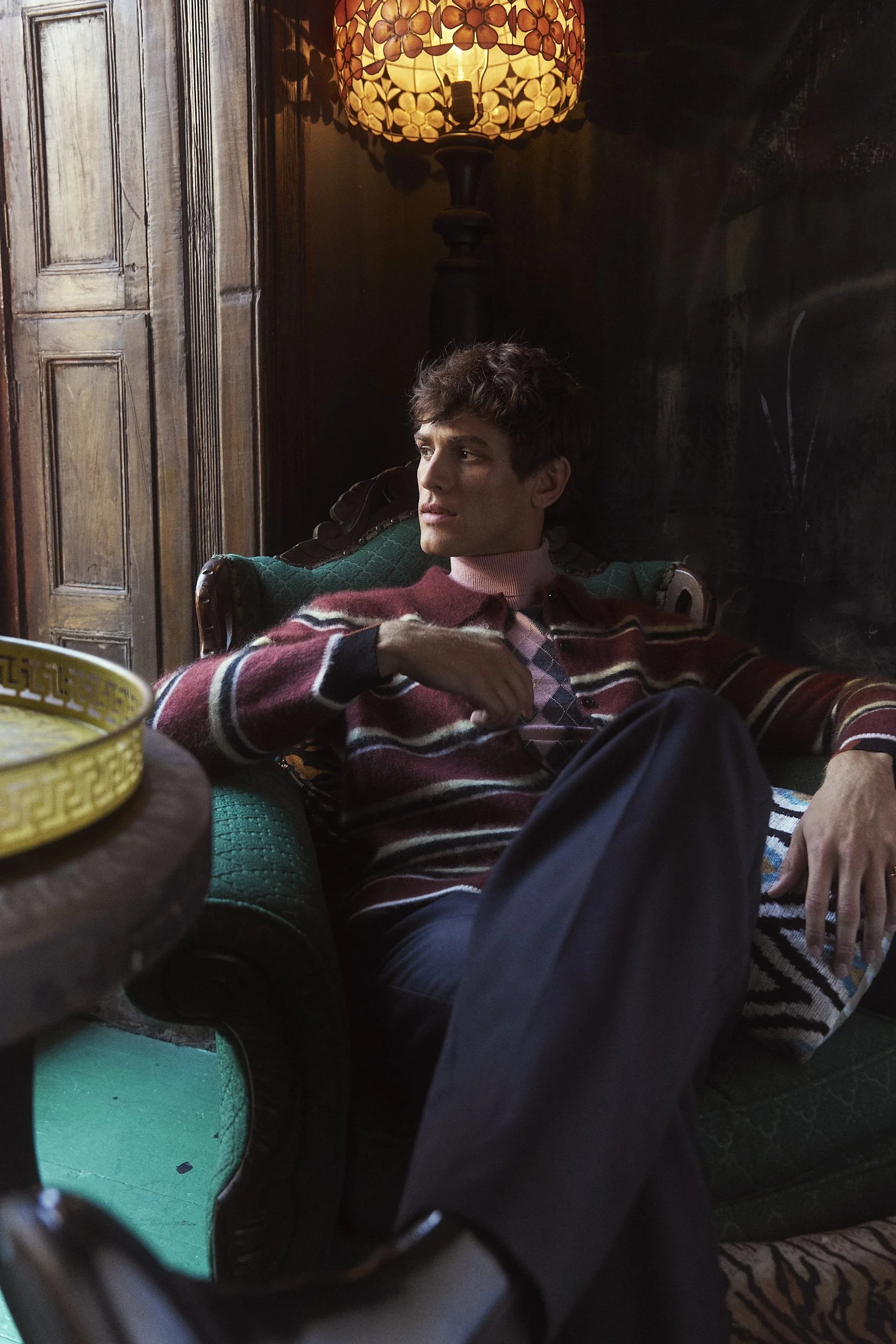 A young man with brown hair sitting on a vintage green sofa near a wooden wall, illuminated by a floral lamp, wearing a plaid shirt under a multicolored striped sweater.