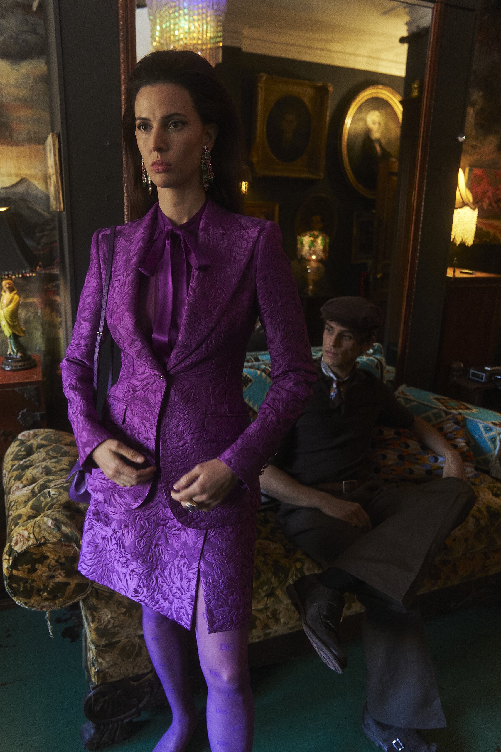 A woman in a purple vintage-style suit with matching tights and large earrings stands in a dimly lit room with decorated walls and vintage portraits. Behind her, a man in a dark shirt, hat, and pants is reclining on a floral-patterned couch with his 