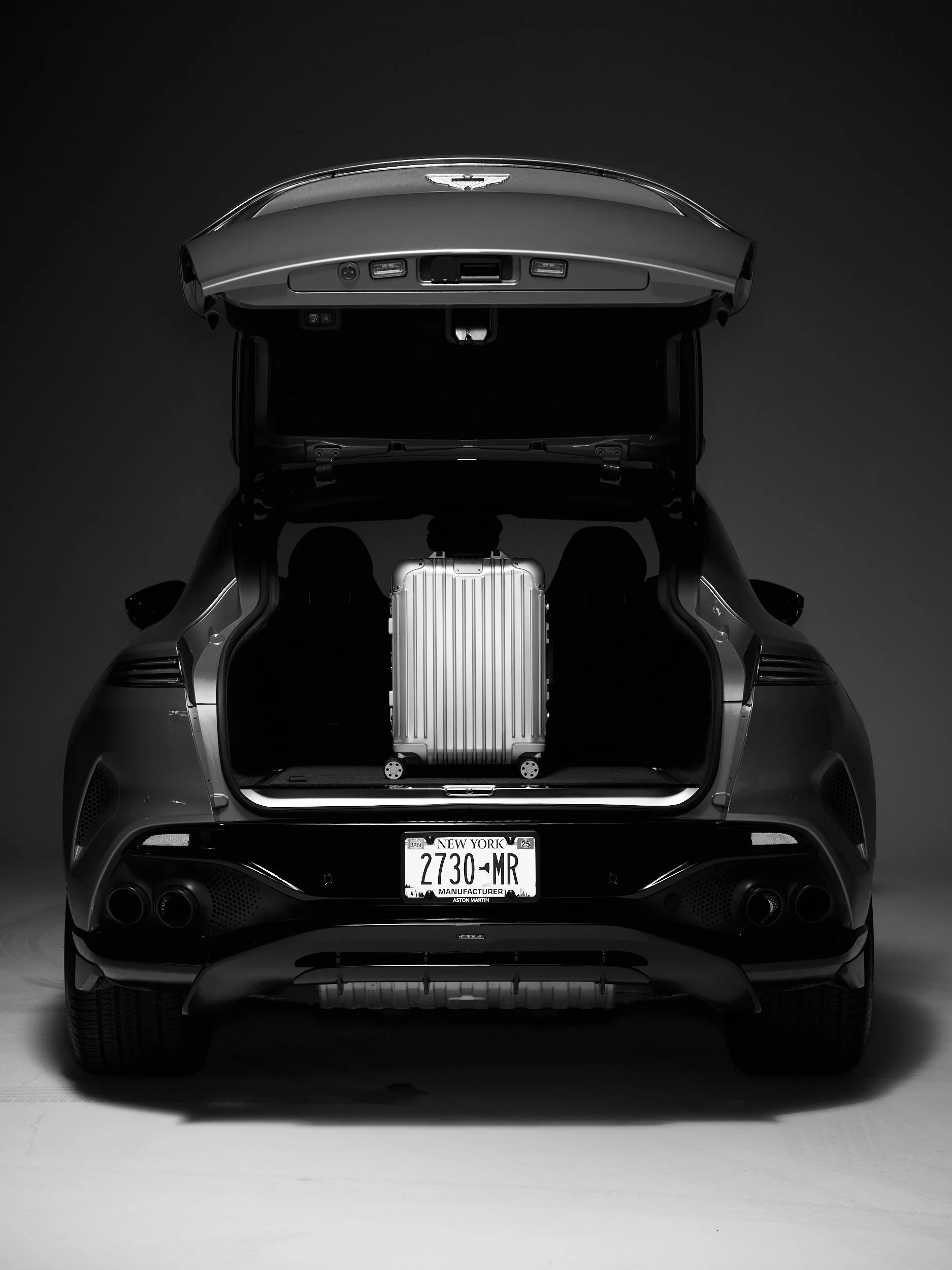 Black luxury car with open trunk, containing a silver suitcase, against a plain dark background.